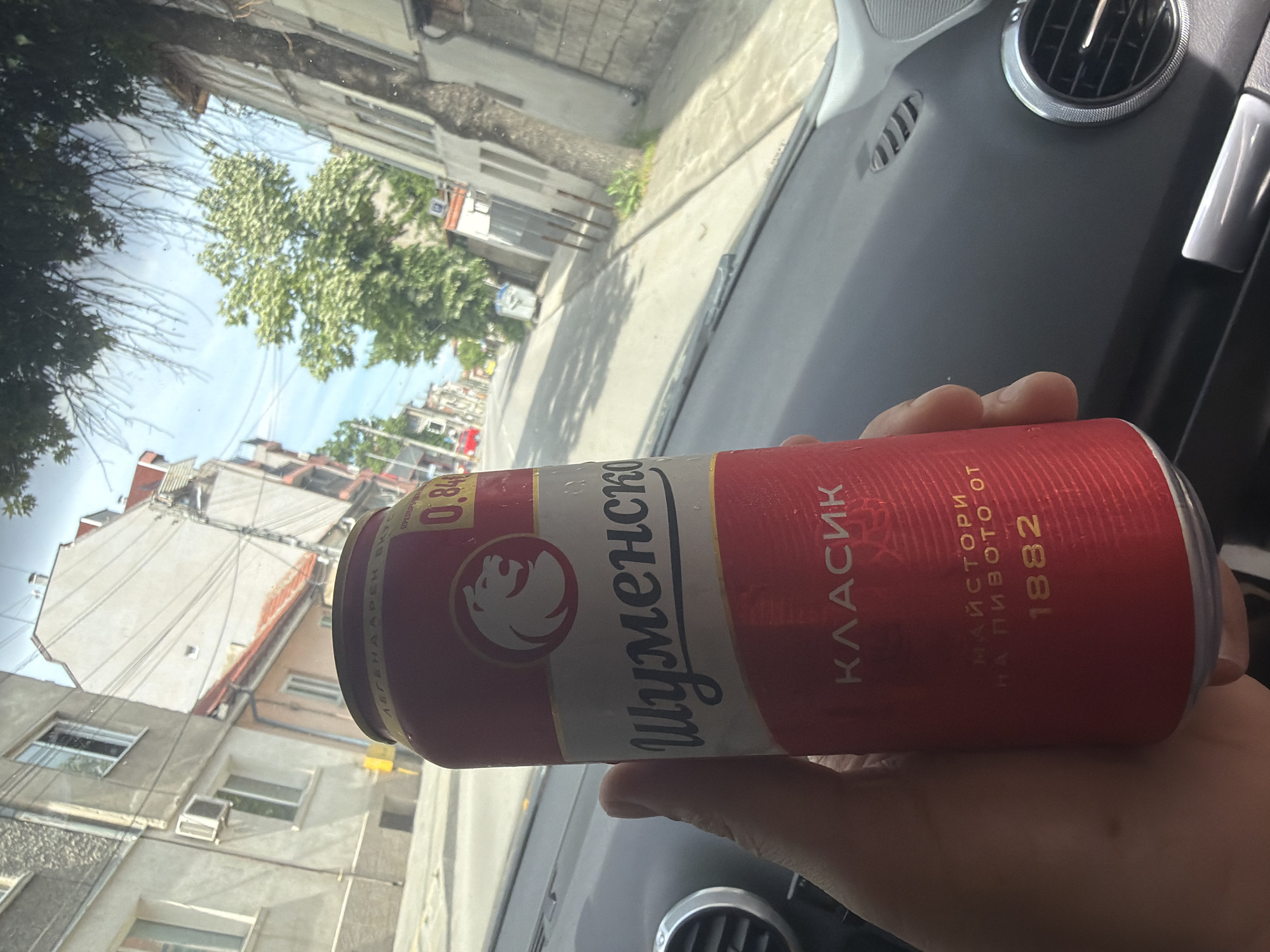 The image presents a first-person perspective from inside a car, looking out onto an urban street scene. The photograph is rotated approximately 90 degrees counter-clockwise.
In the foreground, a hand holds a red and white beer can. The can prominently displays the brand name "Шуменско" (Shumensko) and the word "КЛАСИК" (Classic) in Cyrillic. Other legible text includes "МАЙСТОРИ НА ПИВОТО ОТ" (Masters of beer from) and the year "1882," indicating the brewery's founding. A number "0.84" is also visible, possibly denoting volume. A stylized eagle or bird logo is featured on the can. Parts of the car's interior, including a dashboard section and a circular air vent, are visible in the bottom right of the frame.
Through the car window, a daytime street scene unfolds under a clear, bright sky. The background shows several multi-story buildings, likely residential or mixed-use, with windows and balconies. Green trees with dense foliage are interspersed along the street, which appears to be paved. Visible streaks or smudges on the car window are noticeable against the sky.
The activity depicted is a person holding a can of beer while inside a car, possibly parked or moving slowly. The bright natural light suggests it is daytime with clear weather. Based on the "Шуменско" beer brand, the location is contextualized as Shumen, Bulgaria.
