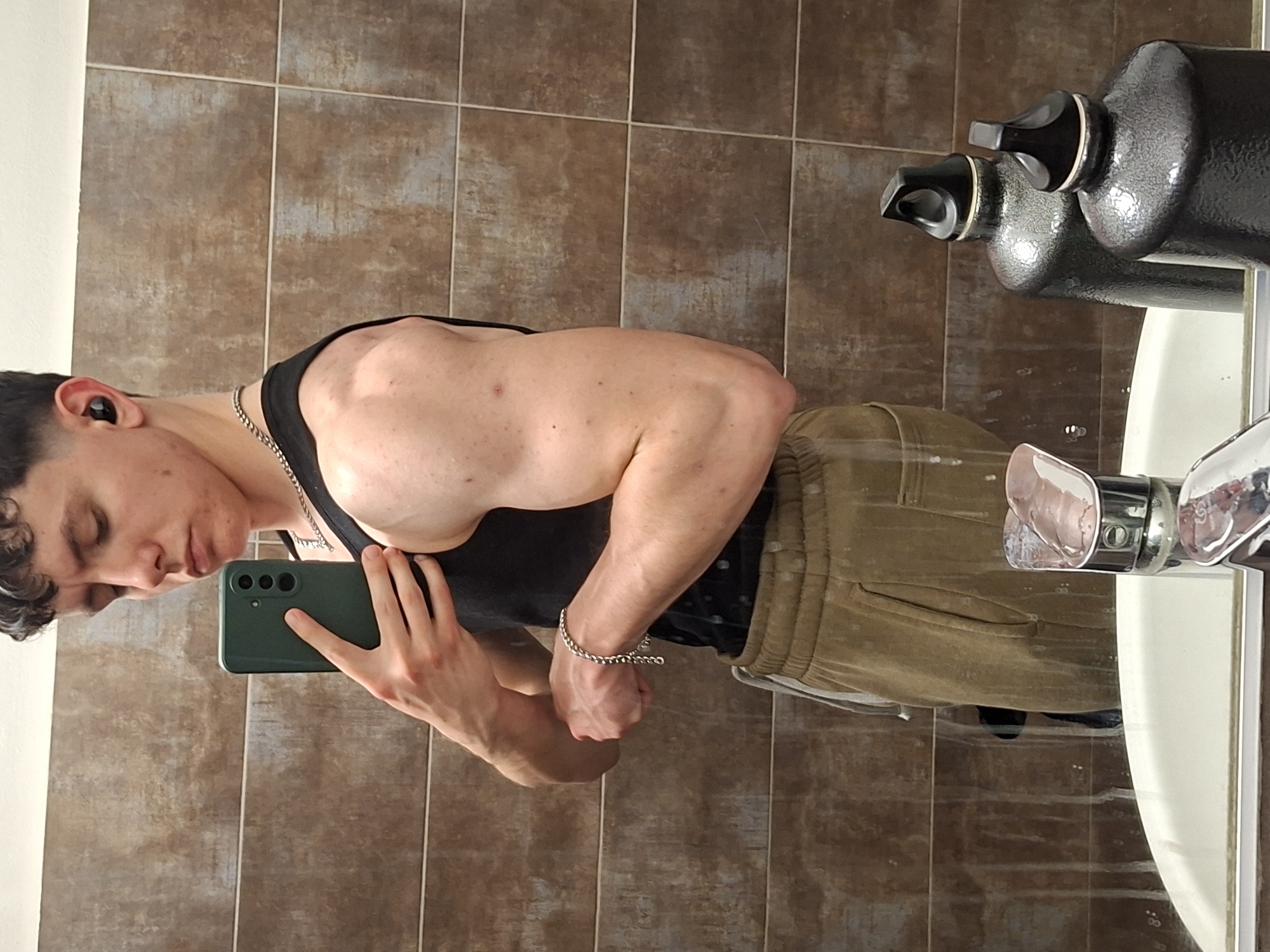 A male is posing in a bathroom mirror, taking a photo with a green smartphone. He is wearing a black tank top, tan sweatpants, a silver necklace, and a silver bracelet. The bathroom has brown tiled walls and a white sink with a metal faucet. Two large black liquid containers are on top of the sink. The location cannot be confirmed to be Plovdiv, Bulgaria.