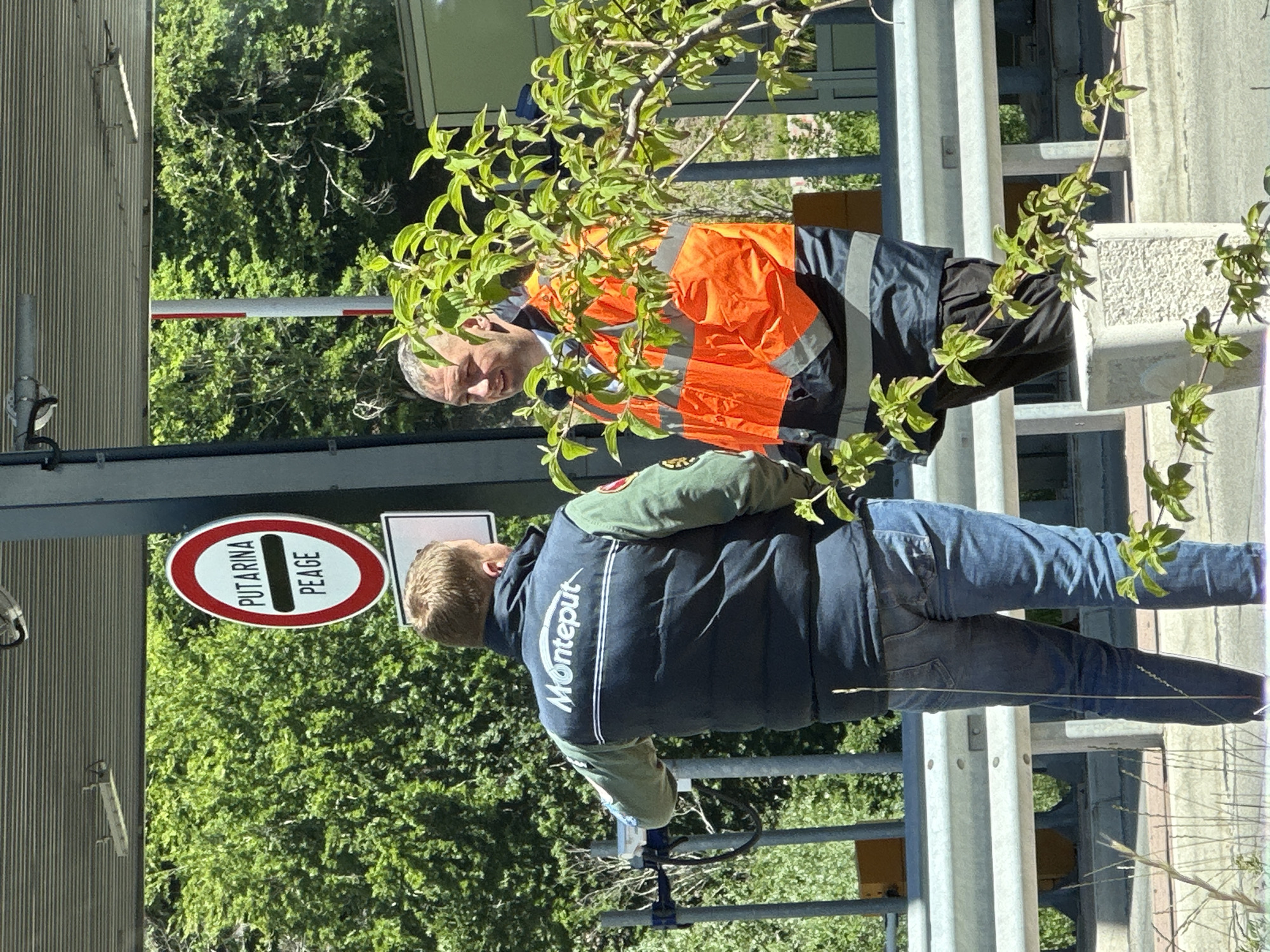 Two men are standing in front of a toll booth structure. The man on the left, with short blond hair, is wearing a dark blue puffy vest over a green long-sleeved shirt and blue jeans. The vest has the white text "Monteput" printed across the back. He is facing away from the camera, looking towards the right. The man on the right, with short gray hair and a shaven head, is wearing a high-visibility orange vest over a dark jacket and dark trousers. He is facing the man on the left, with a slight smile. A red and white circular sign with the text "PUTARINA PEAGE" is visible to the left of the man on the left, mounted on a pole. The background consists of lush green trees and foliage, suggesting an outdoor environment with vegetation. The overall scene appears to be a functional area related to road infrastructure, likely a toll plaza. The presence of the "Monteput" branding on the vest suggests a connection to the Montenegrin national road management company.