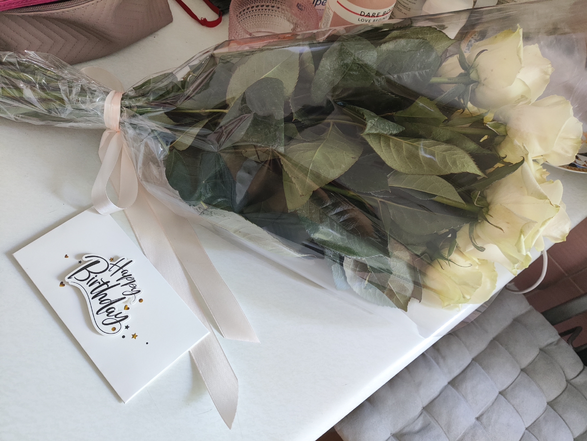 A bouquet of white roses, wrapped in clear plastic and tied with a pale pink ribbon, lies on a white table. A white greeting card with "Happy Birthday" written in black script and decorated with gold stars and hearts is placed next to the bouquet. The scene appears to be indoors.