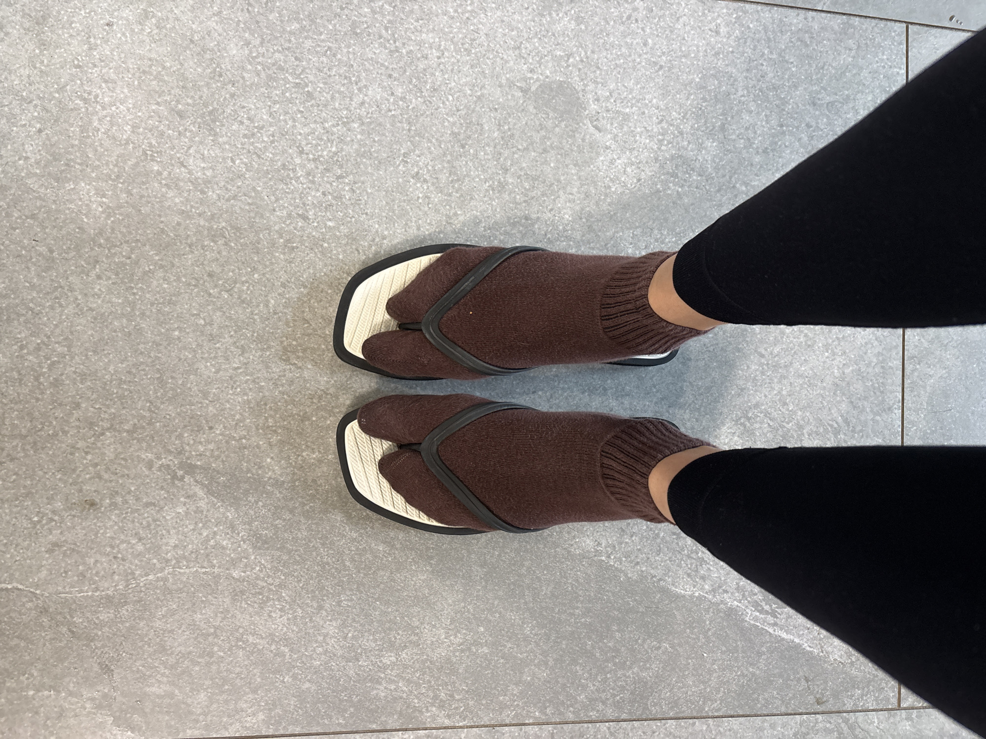 A photograph taken from an elevated perspective shows a person wearing brown tabi socks and geta sandals over a light gray tile floor. The person is also wearing black leggings. The background consists of a solid, light gray, tiled floor with faint lines. There is no indication of location in the city of Chaikovsky, Russia.