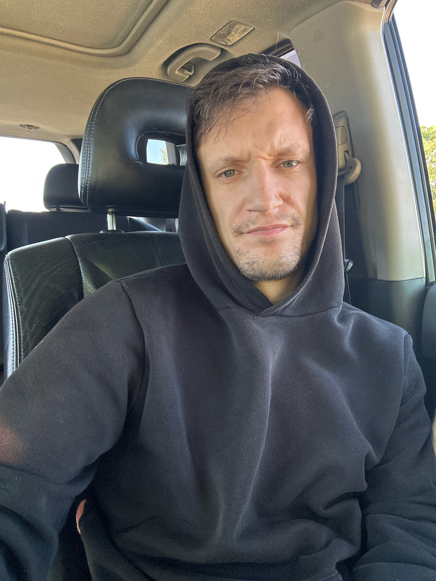 This is a medium shot, eye-level, selfie taken from inside a car. The subject is a man with light brown hair and a faint beard, wearing a black hooded sweatshirt. He is looking directly at the camera with a slightly furrowed brow and a neutral expression. The interior of the car, including the black leather seats and headrests, is visible in the background. The overhead sunroof is also visible. The scene appears to be outdoors, with trees and sunlight visible through the car windows. No specific location within the city of Makiv, Ukraine can be determined from the visual information provided.