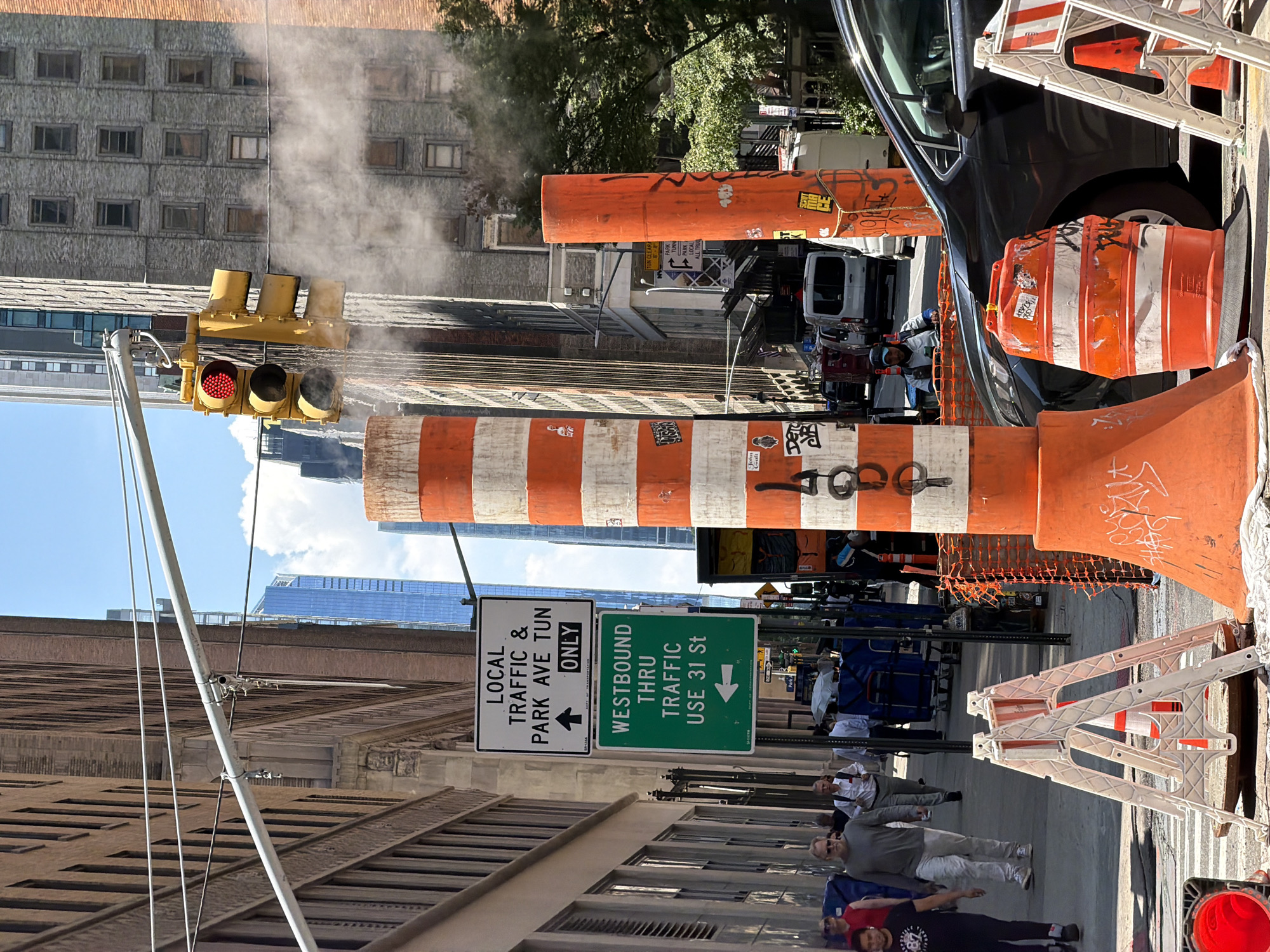In New York City, USA, on a sunny day, construction is underway on a street lined with tall buildings. A traffic light shows red, and steam rises from a nearby utility. Orange and white traffic barrels and cones are placed on the street and sidewalk, indicating a road closure or detour. Several people are walking on the sidewalk, some appearing to be construction workers, while others are pedestrians. Vehicles, including vans and cars, are present on the street. Signs direct local traffic and advise westbound traffic to use 31st Street. The scene suggests an active urban environment undergoing infrastructure work.