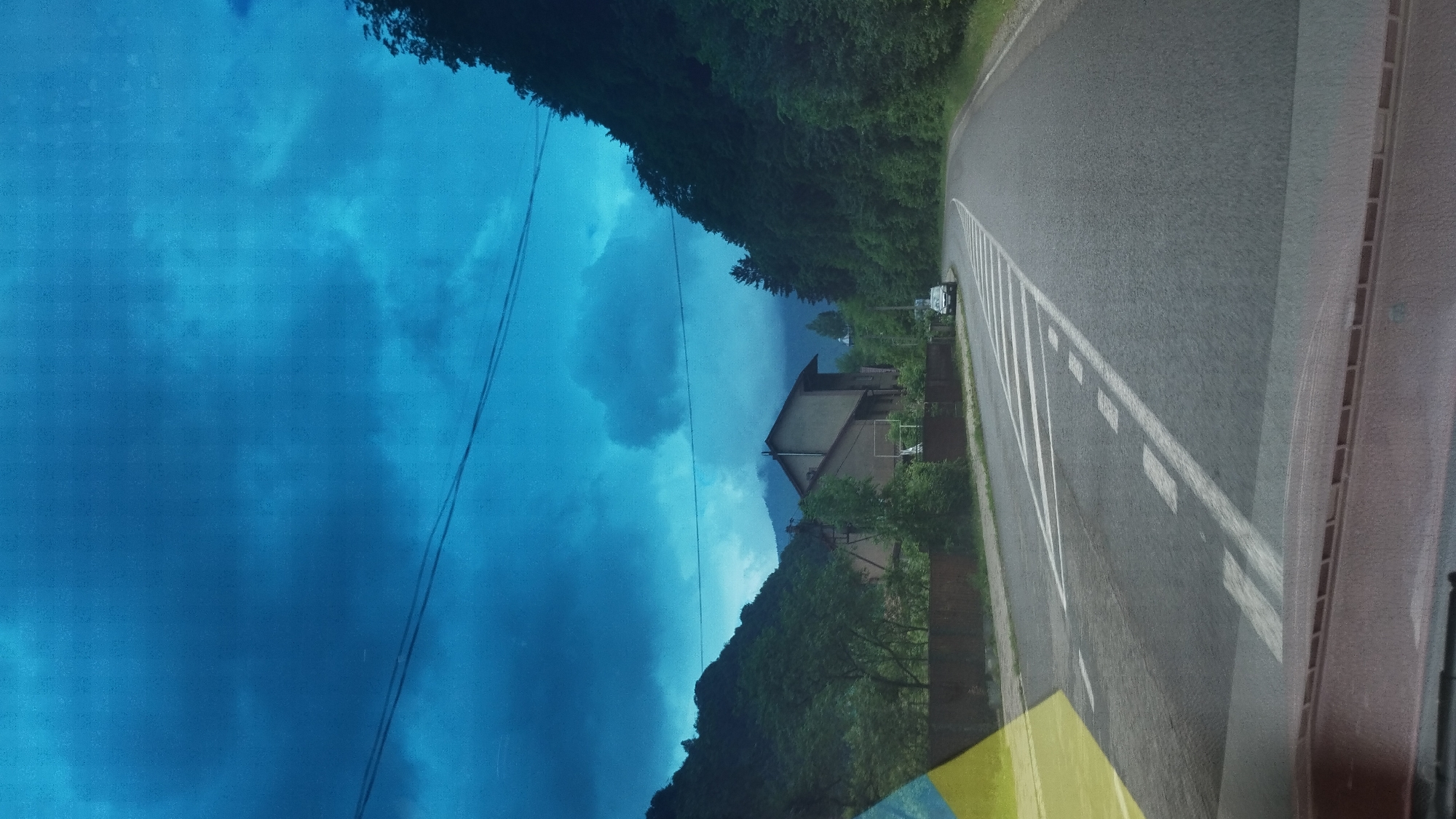 The image depicts a view from inside a vehicle, likely a car, on a road near Rakhiv, Ukraine. The right side of the image shows an asphalt road with white lane markings, bordered by green vegetation. A vehicle is parked alongside the road. On the left side, the view is dominated by a blue sky with dark cloud cover, possibly reflecting in a windshield. A building is visible amidst the greenery, further away from the road. Overhead power lines are also present. The image is slightly rotated, with the road oriented vertically.