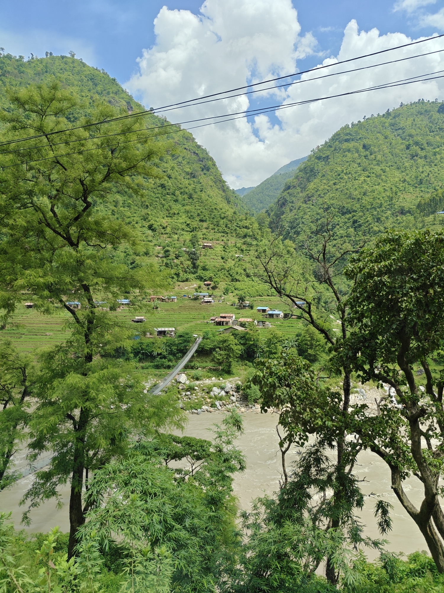 The image depicts a scenic view of a village nestled in a lush green valley in Jogimara, Nepal. A wide, muddy river flows through the valley, spanned by a suspension bridge. The slopes of the surrounding hills are terraced and cultivated, dotted with small houses and scattered trees. The sky is partly cloudy with bright white cumulus clouds against a clear blue expanse. Power lines are visible in the upper portion of the frame, stretching across the landscape. The foreground is dominated by dense green foliage and the branches of trees, partially obscuring the view of the village and the river. The overall impression is one of a tranquil rural environment, characterized by natural beauty and human habitation integrated into the landscape.