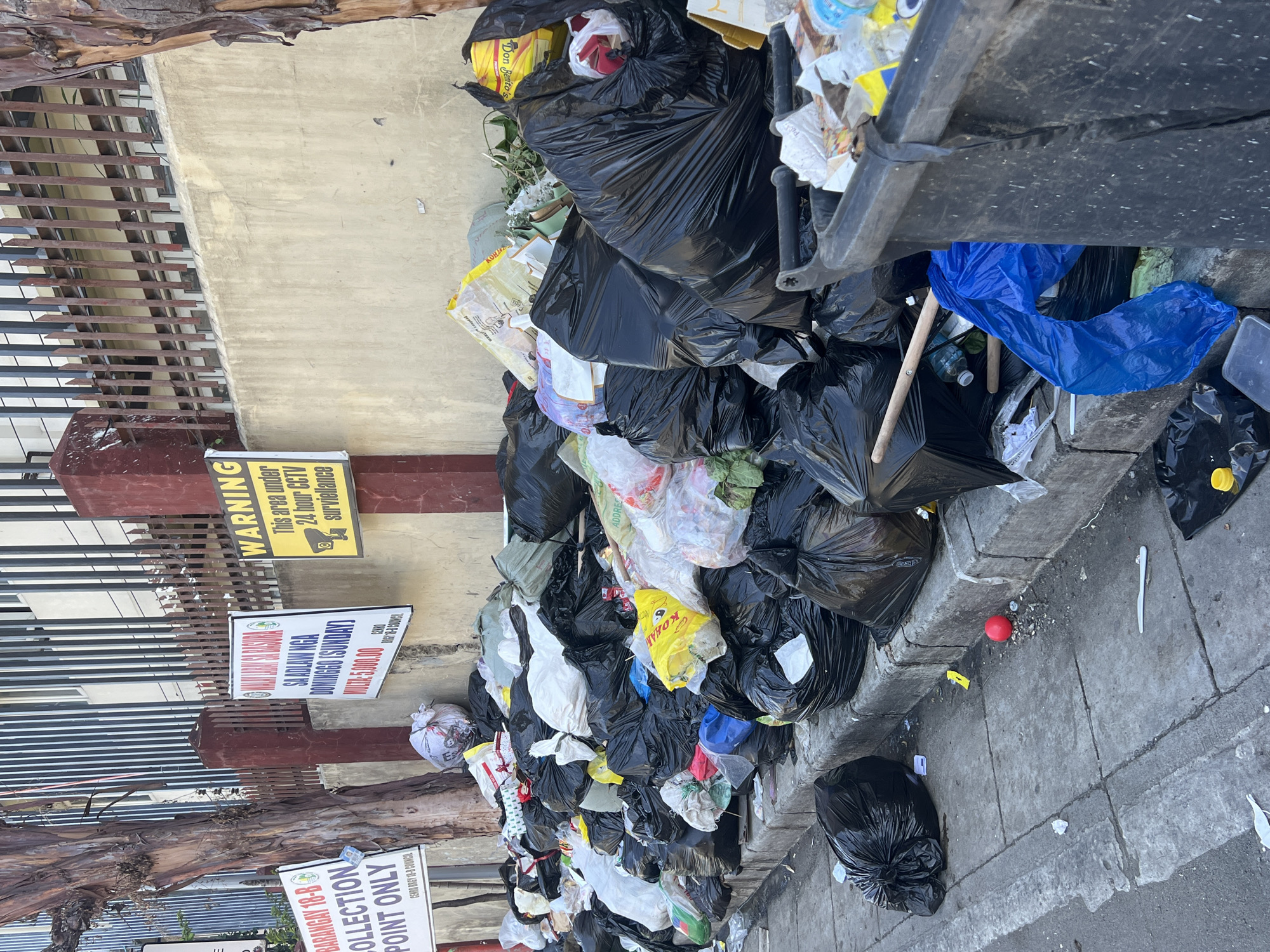 The image shows a large pile of garbage bags and trash containers accumulating on the sidewalk beside a concrete wall and metal fence. Signs posted nearby indicate this is a designated collection point for Barangay 18-B in Davao City, Philippines, and warn of 24-hour CCTV surveillance. A sign also indicates a fine for illegal dumping. The garbage consists mainly of black plastic bags filled with refuse, mixed with visible items such as plastic bottles and food containers.