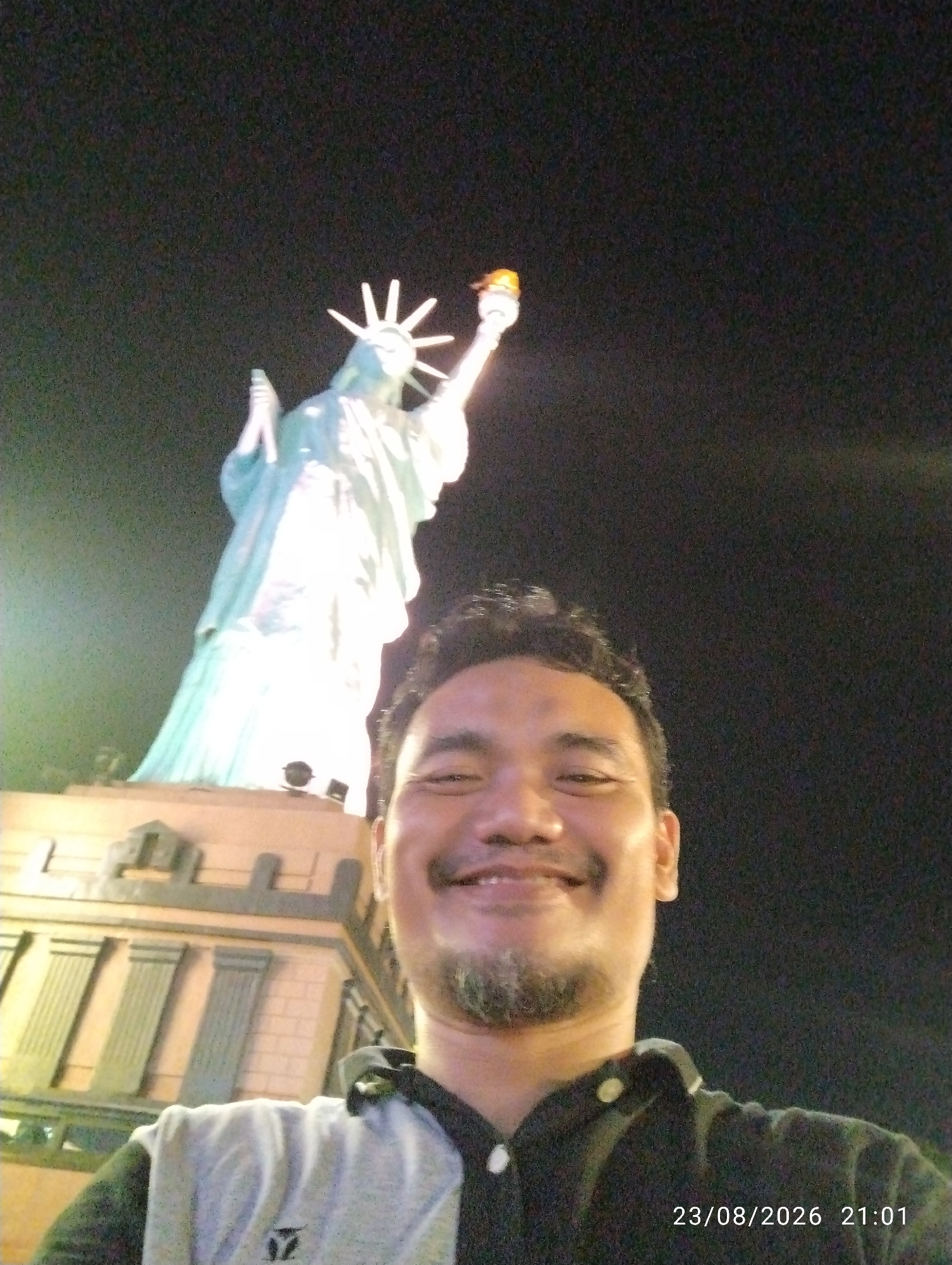 A man is taking a selfie in front of a replica of the Statue of Liberty at night. The man is in the foreground, smiling at the camera and wearing a grey and black collared shirt. Behind him, the Statue of Liberty replica is illuminated and visible against the dark night sky. The base of the statue appears to be part of a building. The timestamp "23/08/2026 21:01" is visible in the lower right corner of the image. This scene is located at the Sasono Langen Kerto amusement park in Madiun, Indonesia.