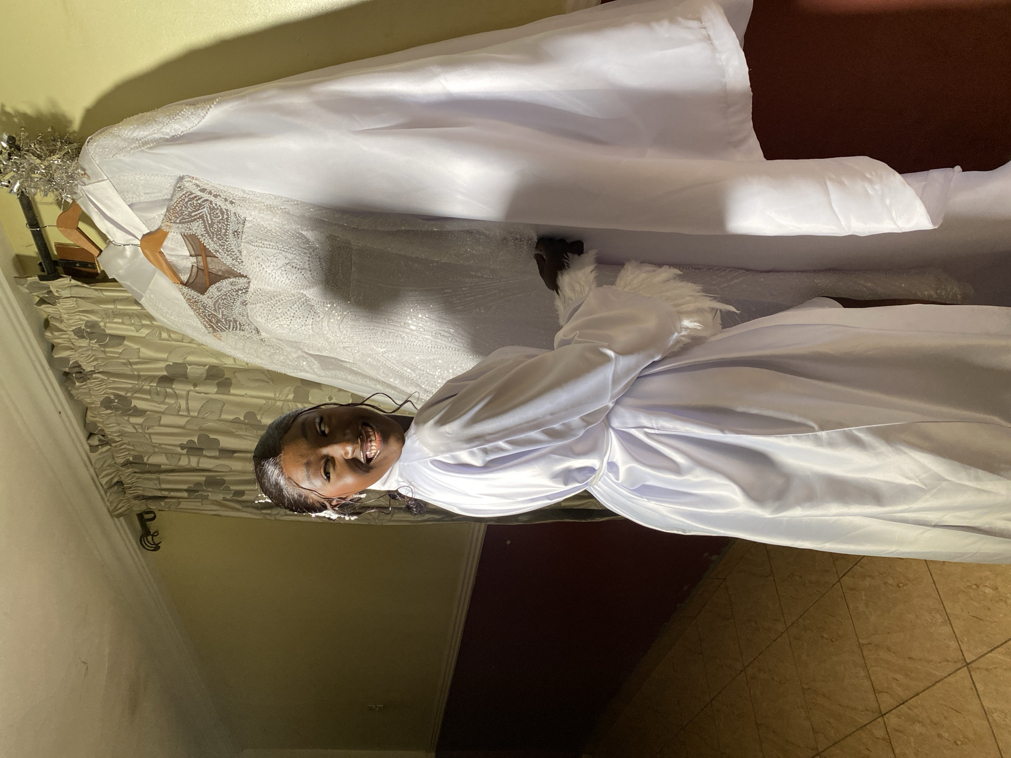 A young Black woman, wearing a white satin robe with feathered cuffs, stands smiling in what appears to be a room. A wedding dress hangs on a hanger, partially obscured by a long white satin fabric draped over it. The room has tan-colored walls, patterned curtains, and tiled flooring. The location is given as Barikin Sabon Gidan Kanat, Nigeria.