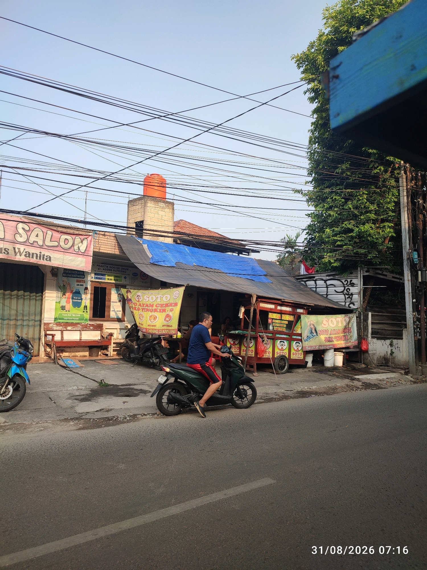 This image depicts a street scene in East Jakarta, Indonesia, on August 31, 2026, at 07:16 AM. The foreground shows a paved road with a motorcycle carrying a person in the middle lane, moving to the right. To the left of the road is a sidewalk area with several parked motorcycles and a blue one prominently featured in the lower left.
Adjacent to the sidewalk is a row of buildings. The leftmost building has a banner that reads "SALON" with smaller text below it. To the right of the salon is a structure with a blue tarp covering its roof. Underneath this tarp, there are food stalls or small shops. One stall displays banners advertising "SOTO AYAM CEKER" and "SOTO AYAM SEGER". Another stall has a red cart with multiple circular signs depicting a cartoon face, and the word "SOTO" repeated.
A person is seen riding a dark green motorcycle in the middle of the frame, heading towards the right. They are wearing red shorts and a blue shirt. Behind the food stalls, there is a tall, brick structure topped with an orange water tank. The environment is characterized by a clear sky with an abundance of overhead electrical wires crisscrossing the scene. A large tree with lush green foliage is visible on the right side of the image, partially obscuring a building. The overall impression is a busy street with commercial activity and residential elements.