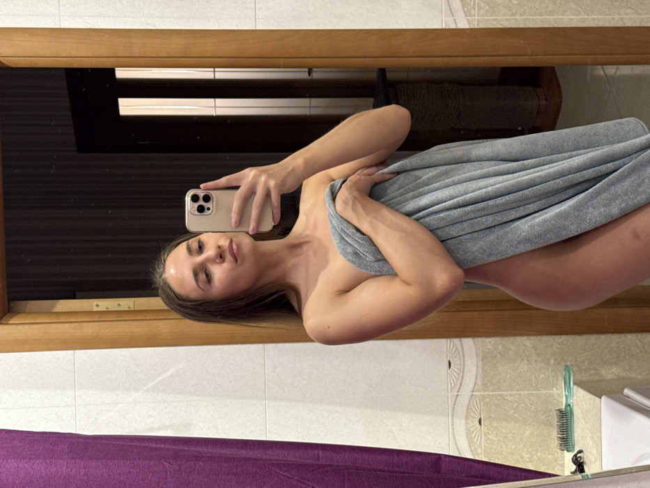 A young adult female is depicted in a mirror selfie taken inside what appears to be a bathroom. She has long brown hair and is partially nude, holding a light grey towel wrapped around her torso and upper thighs with her left hand. Her right hand holds a light-colored smartphone with a clear case, positioned to capture the image. Her expression is neutral, with her gaze directed towards the phone. To her left, a purple fabric shower curtain is visible, along with white tiled walls featuring a decorative wave-patterned border tile. A green-handled comb or brush rests on a white shelf or ledge in the lower left foreground. To her right, a light brown wooden door frame is reflected in the mirror, revealing an adjacent room with dark, possibly vertically striped walls, and a multi-level cat scratching post. The floor appears to be tiled with light beige rectangular tiles. The precise location within Saint Petersburg, Russia, is not discernible from the visual information provided, but the scene indicates a private residential interior.