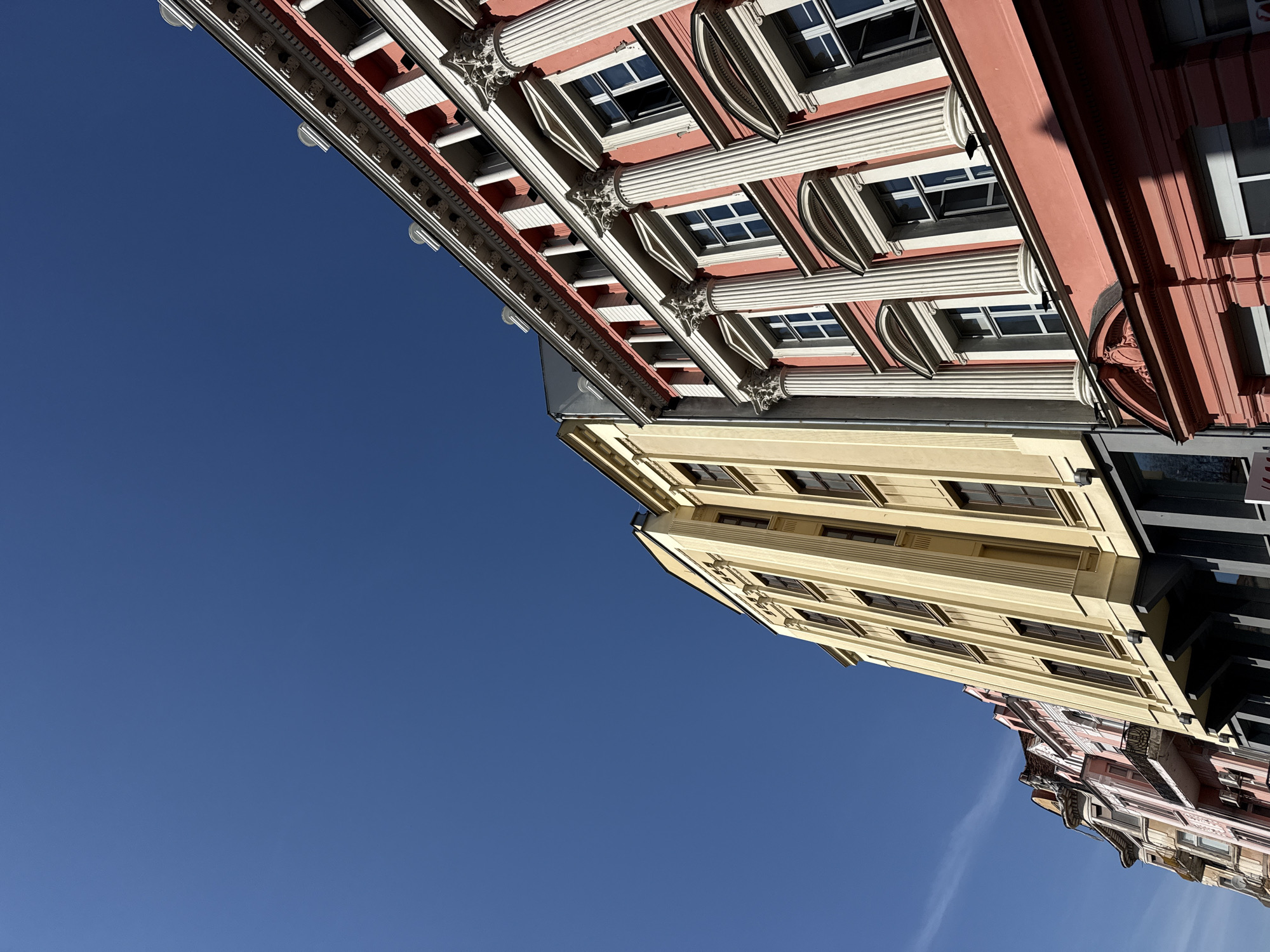 The image features a low-angle shot of colorful buildings in Plovdiv, Bulgaria, against a clear blue sky. The buildings are of different colors including red, yellow, and pink. The buildings feature architectural details like columns, windows, and decorative trim. The composition is such that the buildings appear to converge in the distance.