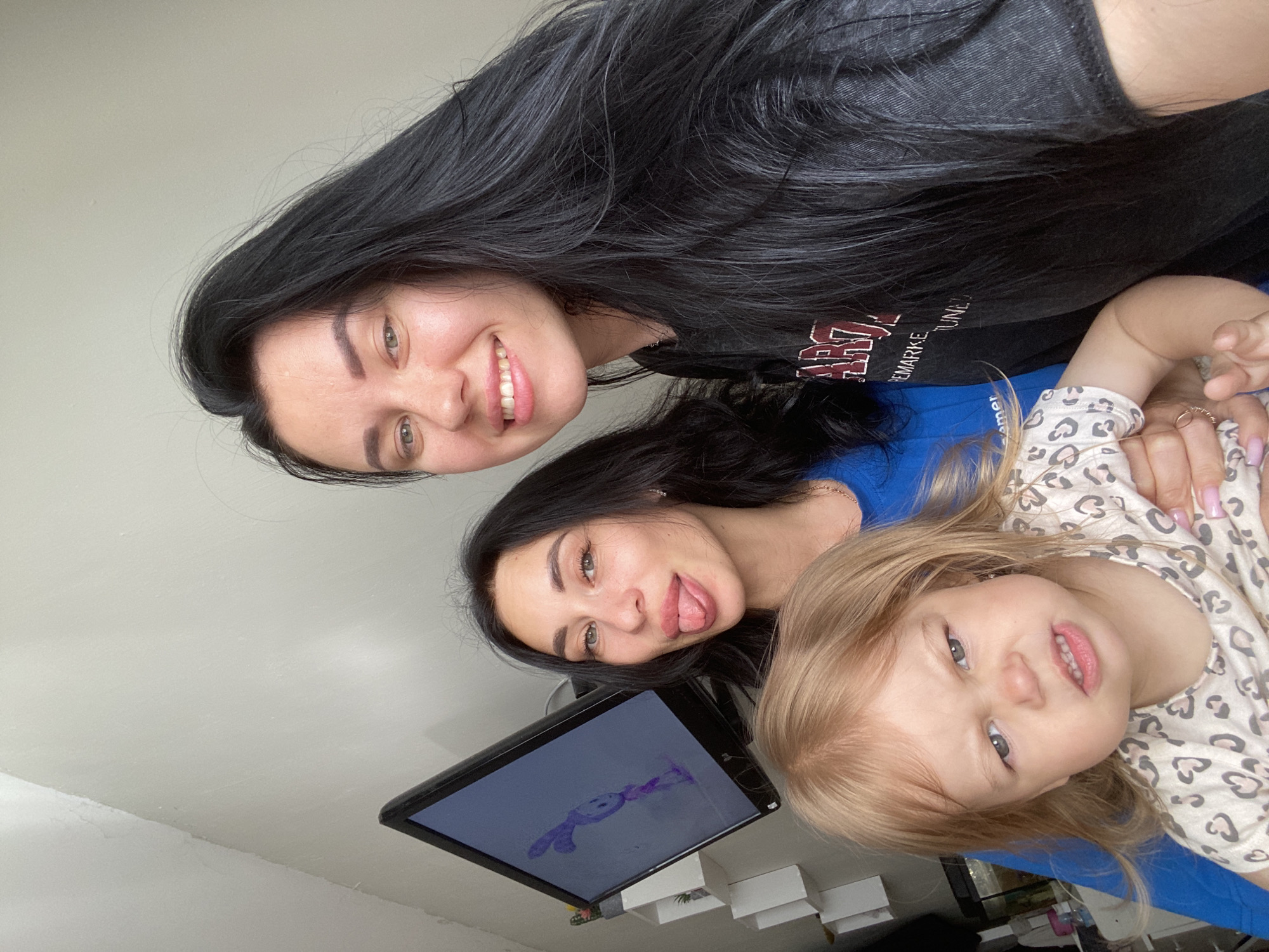 A group of three people are captured in a selfie-style photograph. In the foreground, a young Caucasian female child with blonde hair and a pouting expression is held by an unseen person. The child wears a light-colored top with a leopard print pattern. Immediately behind the child, a Caucasian female with long dark hair and light-colored eyes is positioned to the left. She is sticking her tongue out playfully. To the right of her, another Caucasian female with very long dark hair and brown eyes smiles broadly at the camera. She is wearing a dark gray t-shirt with text that is partially visible. The background consists of a plain white wall and a computer monitor displaying a cartoon character. A shelf unit with white cubbies is visible to the left of the monitor. The overall setting appears to be indoors, likely a residential environment. The image does not contain enough information to determine the specific location within the city of Chaikovsky, Russia.
