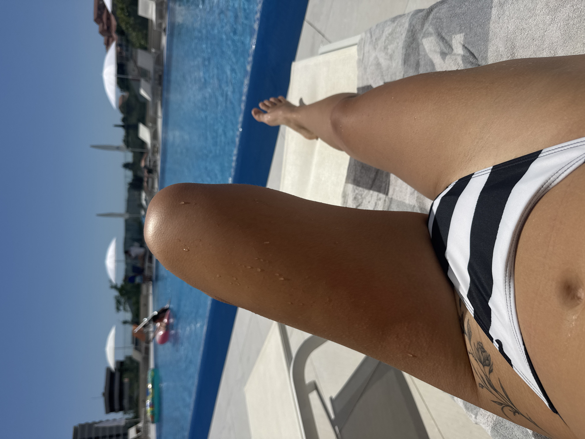 The image depicts a first-person perspective of a person relaxing on a sun lounger at a swimming pool in Boreti, Montenegro. The foreground shows the tanned lower torso, upper thighs, and part of the left arm of an individual, adorned with a black and white striped bikini bottom. Small water droplets are visible on the skin. A black floral or vine-like tattoo extends from the upper left thigh towards the hip. The person is situated on a white sun lounger partially covered by a grey and white striped towel.
In the midground, a large, clear blue swimming pool occupies the center of the frame, with a distinct blue tiled edge. Several white sun loungers and white parasols are positioned around the pool. The pool contains inflatable objects, including a colorful ring and a pink float, and blurred figures of people are visible either in the water or beside it.
The background reveals a bright, cloudless blue sky, indicating clear weather. Architectural structures, including a multi-story building on the left and buildings with reddish roofs on the right, are visible in the distance, interspersed with greenery.
