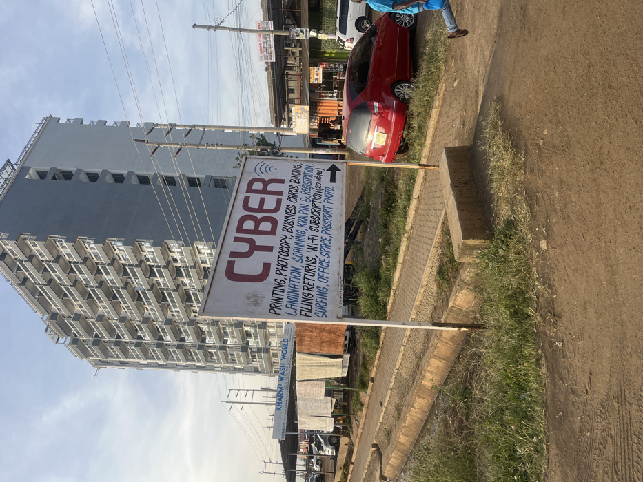 A sign advertises "CYBER" services including printing, photocopying, business cards, binding, lamination, scanning, KRA PIN & registration, filing returns, Wi-Fi subscription (20 Mbps), surfing, office space, and passport photos. Another sign indicates "KHARM WASH WORLD SERVICES" is nearby. In the background, a tall, modern building rises behind power lines, and cars are parked on the street. The image appears to be taken on a sunny day.
