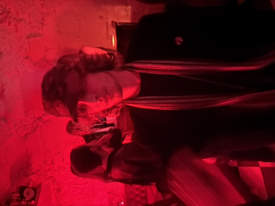 The image depicts an interior scene within a subterranean establishment, likely a nightclub or bar, characterized by rough-hewn, stone-like walls bathed in intense red illumination. This architectural style, featuring vaulted or cave-like structures, is common in repurposed historic cellars found throughout Paris, France.
In the center-right foreground, a young male subject is positioned, facing slightly towards the camera. He wears a dark cap, obscuring his eyes with shadow, and has medium-length, wavy dark hair. His attire consists of a dark jacket or coat over a dark shirt, accessorized with a vertically striped scarf, predominantly red and light-colored, draped around his neck. A small, circular pin or badge is affixed to the right lapel of his jacket. His expression is neutral with slightly parted lips.
In the out-of-focus background, several other individuals are present, standing and interacting, typical of a social gathering in a crowded venue. A padded, possibly quilted, wall section is visible on the far left. A faint checkered pattern is discernible on the floor in the lower-left quadrant. High on the left wall, a speaker or light fixture is mounted. The scene conveys an atmospheric, active social environment within a distinctively Parisian underground venue.