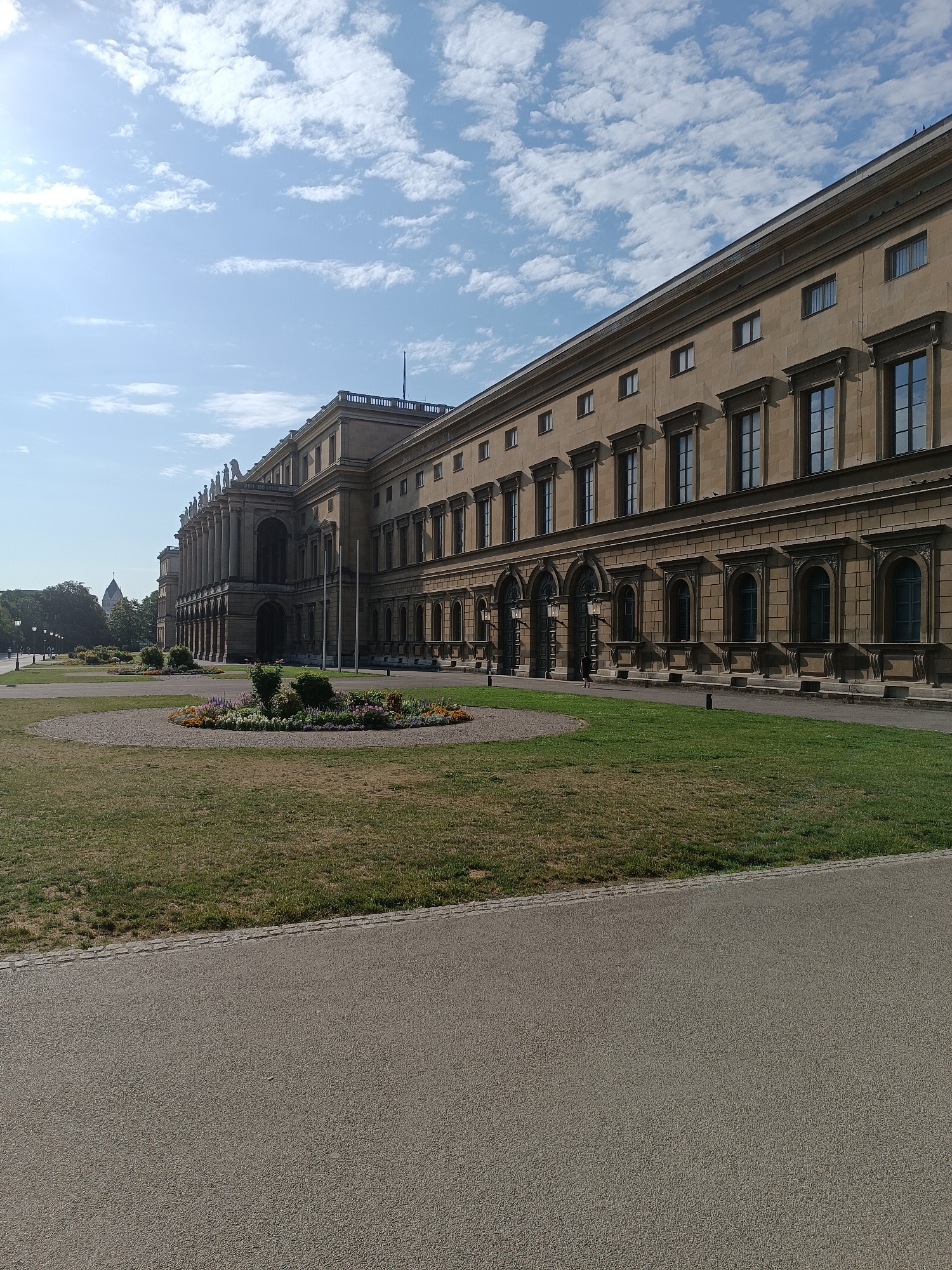 The image depicts the west wing of the Residenz in Munich, Germany, bathed in sunlight. The grand building, constructed from tan stone, features numerous arched windows and decorative moldings. A long row of stately columns lines the facade, and classical sculptures adorn the roofline. In the foreground, a well-maintained lawn leads to a circular flowerbed filled with colorful blooms. Beyond the building, a few trees are visible, and in the distance, a dome-shaped structure can be seen. The sky is a clear blue, dotted with fluffy white clouds, and the sun casts a warm glow on the scene. The overall atmosphere is one of stately elegance and tranquil beauty.