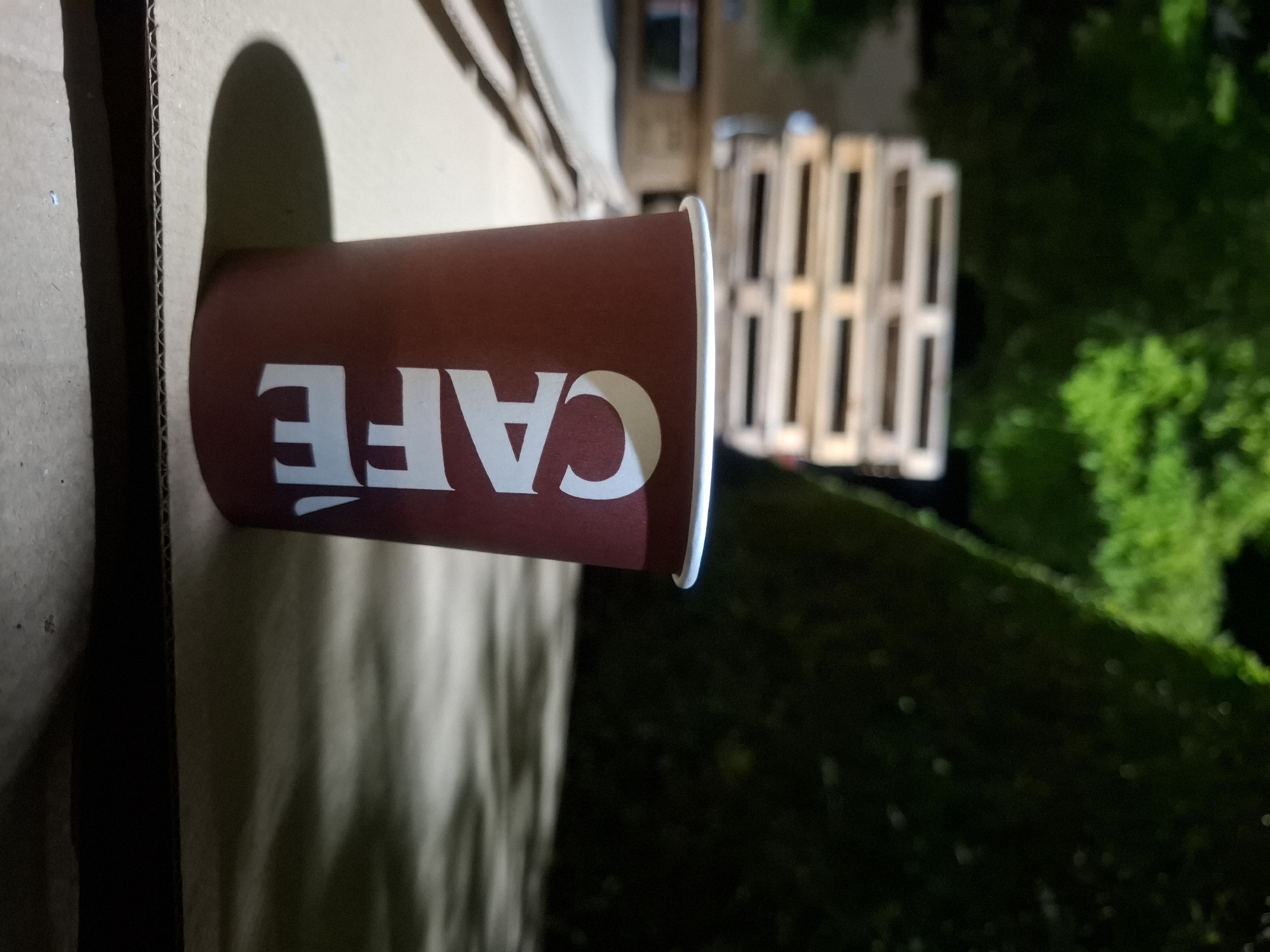 A maroon paper cup with white lettering that reads "CAFÉ" is placed on a cardboard surface. In the background, a stack of wooden pallets is visible, partially obscured by greenery. The scene is dimly lit, suggesting it is nighttime. The image appears to be taken outdoors, possibly in a loading area or an industrial setting, with elements that could be found in various urban locations. No specific identifiable landmarks or context place this image in Plovdiv, Bulgaria.