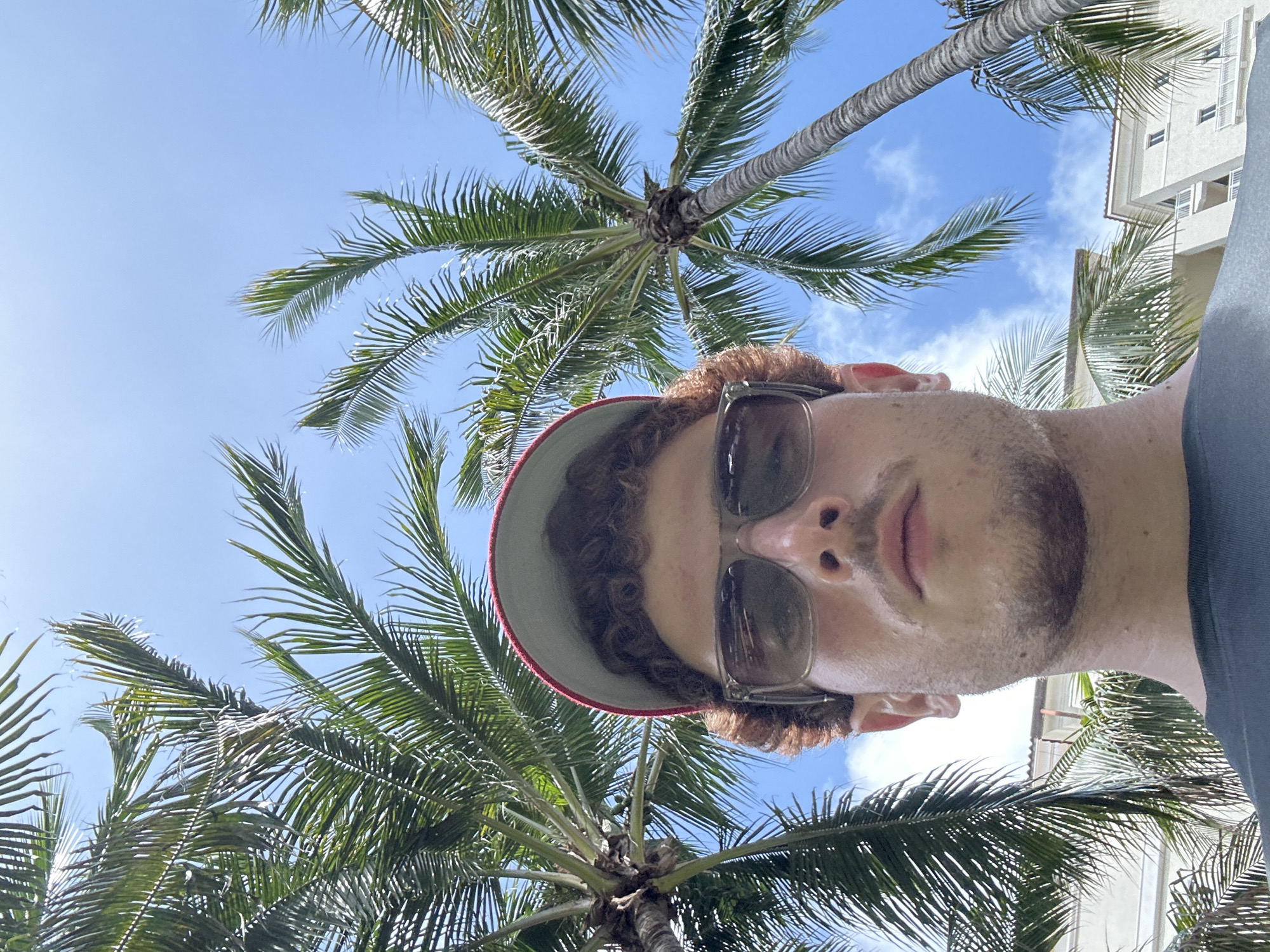 A selfie taken from a low angle under palm trees against a blue sky with some clouds. A man is wearing sunglasses and a hat with a red brim. A building is visible in the background to the right. The image appears to be taken in Aventura, Florida, United States of America, based on the prevalence of palm trees and architectural style, common in the area.