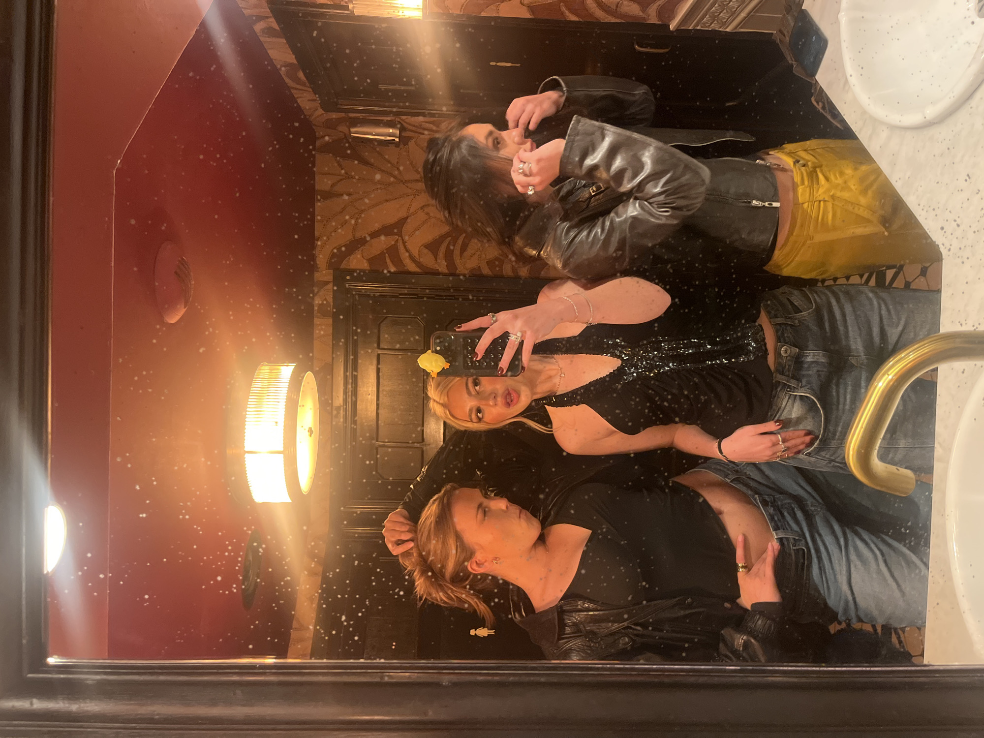 Three women are reflected in a mirror in what appears to be a restroom. The woman closest to the mirror is wearing a black top and blue jeans, and a black jacket is draped over her. The woman in the middle is taking a photo using her phone and is wearing a black sequined top and blue jeans. The woman farthest away is wearing a black leather jacket and yellow pants. There is a sink visible to the right side of the mirror. The restroom's walls are dark red and have a patterned wallpaper. A light fixture is visible on the left side of the mirror. Based on the restroom's interior design and general atmosphere, it is highly probable the location of this photo is in Paris, France, specifically at a trendy or upscale establishment.