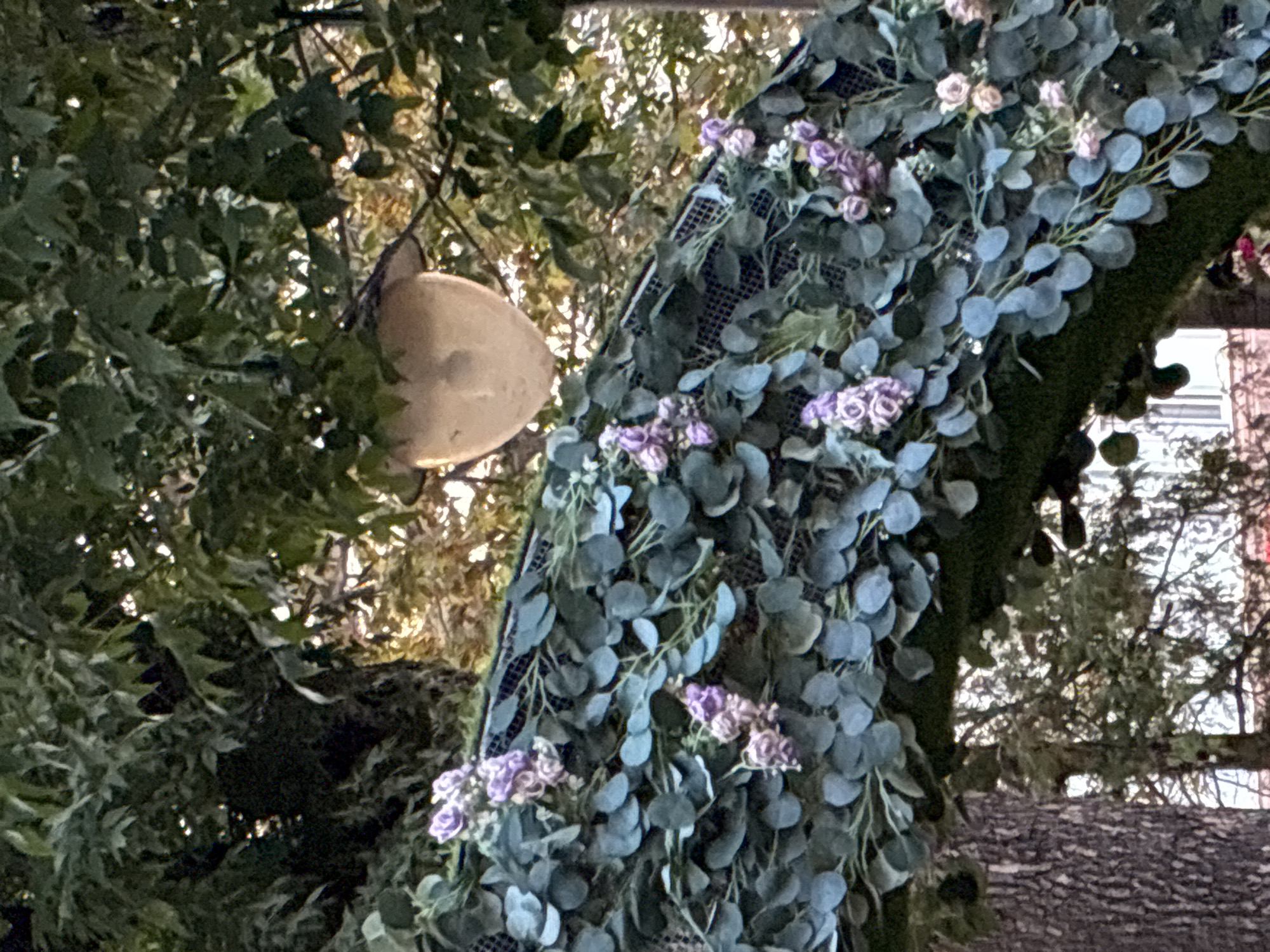 The image captures an outdoor scene, possibly in Mahilyow, Belarus, focusing on a decorative archway. The arch is adorned with artificial green foliage and clusters of small, purple and pink flowers. Hanging from the arch, or from a nearby branch, is a light-colored, spherical object, possibly a lantern or a decorative element. In the background, trees with dense green leaves are visible, with sunlight filtering through them, creating a dappled effect. The lower portion of the image shows the textured bark of a tree on the left and hints of buildings or structures in the blurred background on the right. There are no people visible in the image.