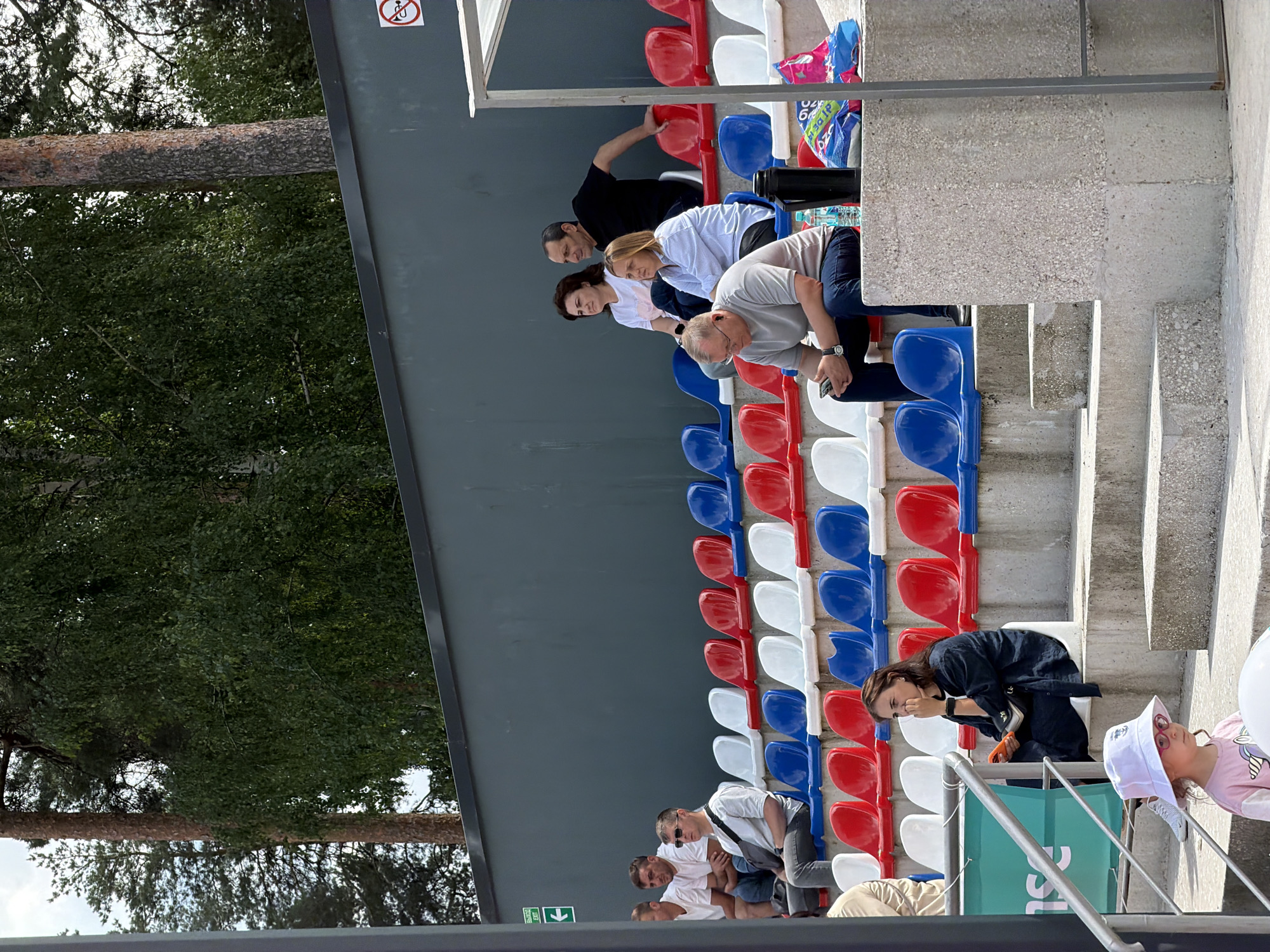 In Bykovo, Russia, a group of people are seated in tiered stadium seating. The seats are a mix of red, white, and blue. Behind the seating area is a dark grey wall with a white sign indicating "EXIT" with an arrow. Trees are visible in the background, suggesting an outdoor setting.
In the foreground, a young girl wearing a white hat and pink shirt with glasses is looking down. To her right, a woman in a dark blue outfit is seated and holding an orange object. Further up the stairs, a man in a grey shirt and dark pants is looking at his phone. Other spectators are seated in the rows above. A black thermos and a plastic bag are on a concrete ledge. The overall scene appears to be a sporting event or outdoor gathering.