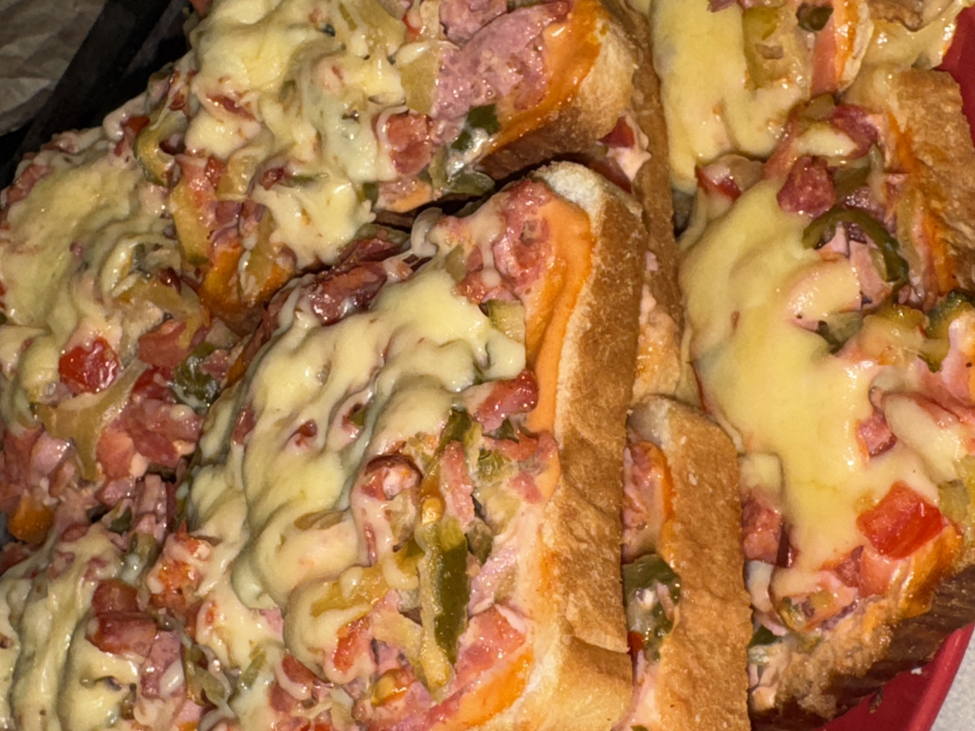The image shows a close-up of food, specifically bread slices topped with a mixture of what appears to be processed meat (possibly ham or sausage), vegetables (likely tomatoes and green peppers), and melted cheese. The dish has a somewhat messy appearance, with the topping overflowing onto the plate. There is no indication of the image's origin in Chaikovsky, Russia. No people, actions, interactions, or additional environmental details are visible.