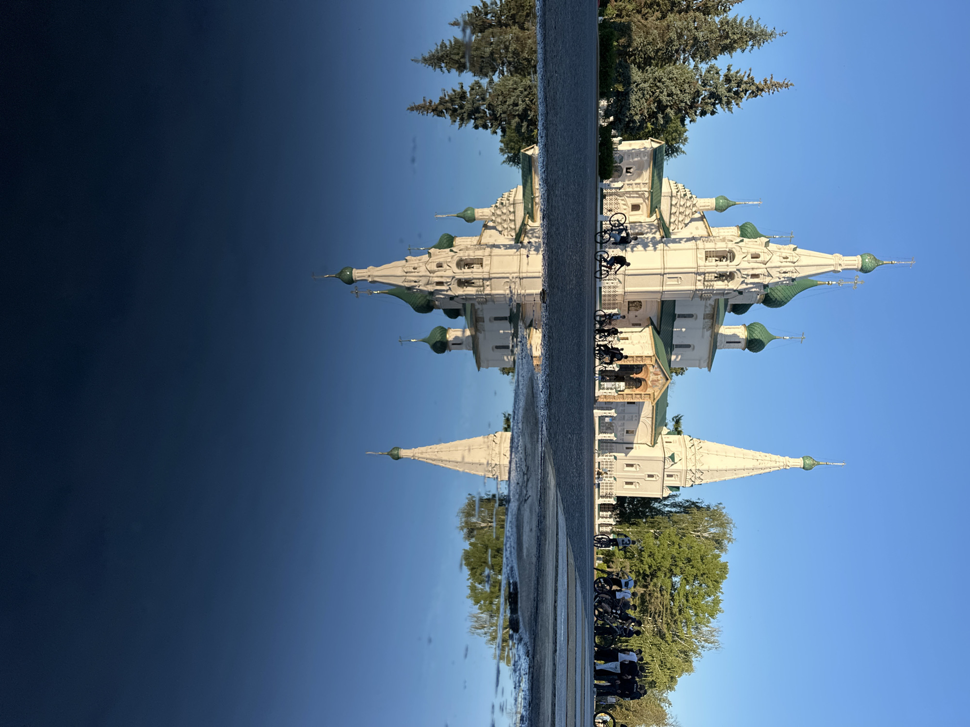 The image depicts a reflection in water, creating an upside-down view of a scene. The primary subject is a white church with green domes, characteristic of Russian Orthodox architecture. It is a sunny day, and the bright blue sky is visible. Trees with lush green foliage surround the church.
In the foreground, which is the reflection, a paved area with some wet patches suggests recent rain or a damp surface. Several bicycles are parked in a row. A group of people, mostly dressed in casual attire, are visible near the bicycles and the church. Their presence suggests a public space, possibly a tourist attraction or a gathering point.
The reflection creates a striking visual effect, with the church and trees appearing to float in the dark, reflective surface which dominates the left side of the image. The light conditions suggest it is daytime, likely morning or afternoon, given the clear sky and bright lighting. The overall mood is serene, with the reflection adding a touch of artistic or dreamlike quality to the scene. There is no visible text in the image. The setting is consistent with a location in Yazvitsevo, Russia, given the architectural style and natural environment.