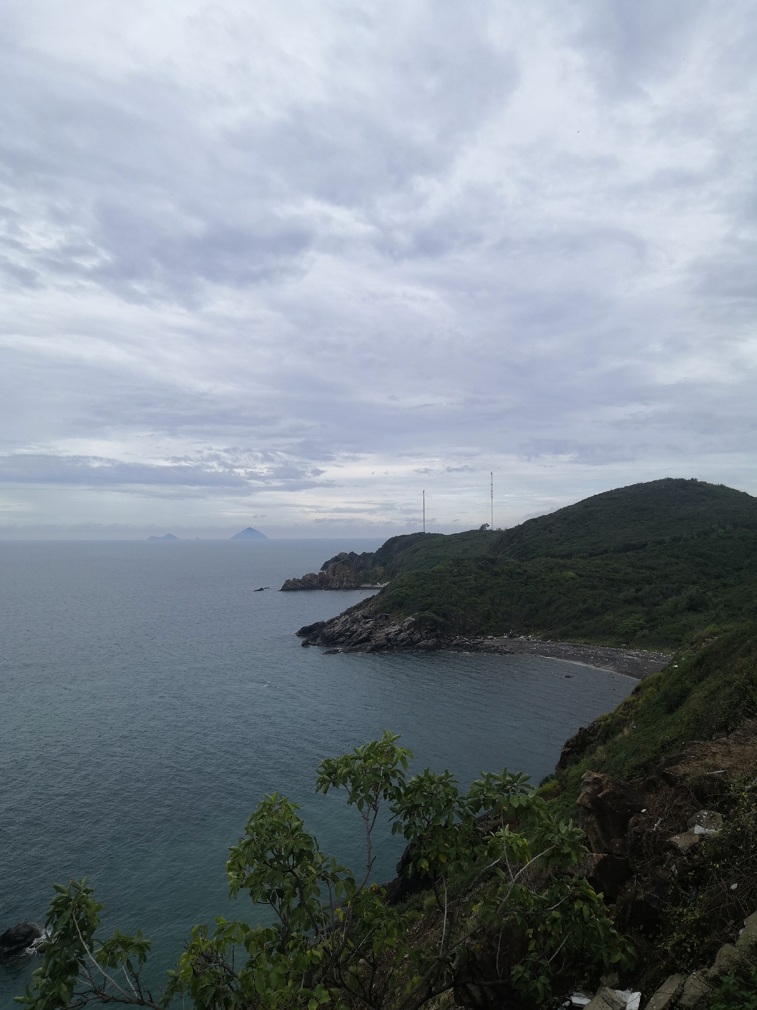 The scene is a coastal landscape in Bac Nha Trang Ward, Vietnam, under a cloudy sky. The sea stretches towards the horizon, with two small islands visible in the distance. A rocky coastline leads to a large, green hill covered in vegetation. Two tall antennas are situated on the hilltop. A small beach with dark sand or pebbles is nestled at the base of the hill. In the foreground, some trees with green leaves are partially obscuring the view. Rocks and foliage are visible at the edge of the cliff in the bottom right corner.