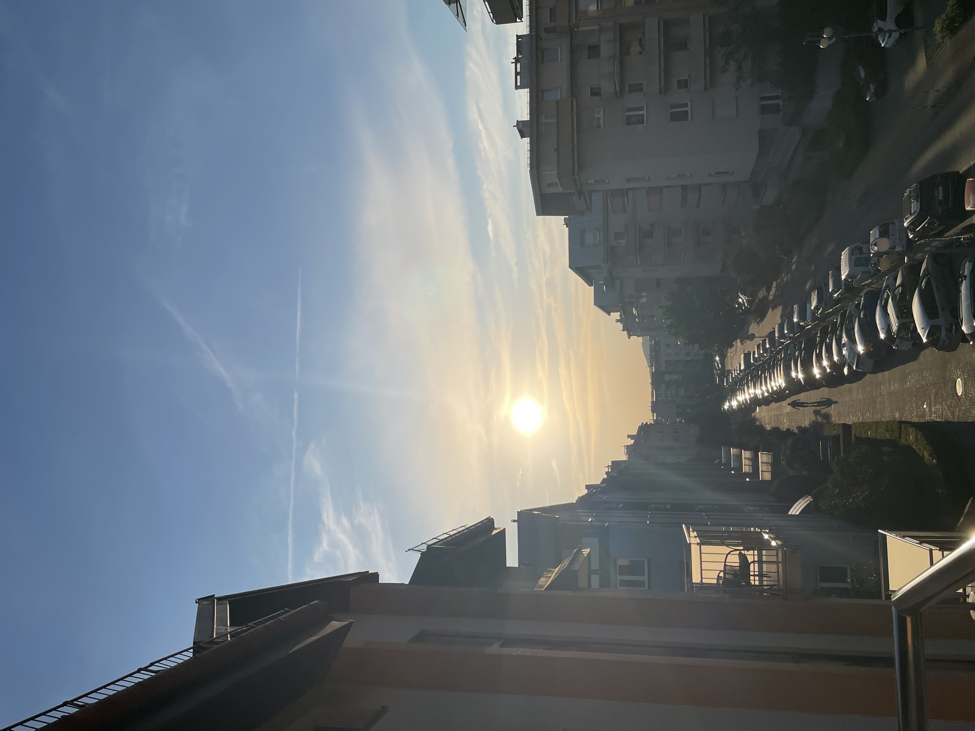An outdoor, vertical, eye-level shot captures a street lined with apartment buildings and parked cars under a bright, hazy sun. The sun, positioned in the upper center of the image, casts a warm glow across the scene, creating lens flare and a soft haze. A few wispy clouds dot the clear blue sky. The buildings, varying in architectural style, rise on both sides of the street, with balconies and windows visible. On the left, a building with a pale orange and white facade is partially in the foreground, featuring a balcony with chairs. On the right, a continuous row of cars is parked along the street, reflecting the sunlight. A single pedestrian walks down the street, casting a long shadow. The street appears to be paved, with some greenery visible between the buildings and along the sidewalk. The overall impression is a tranquil urban setting during a sunny day.