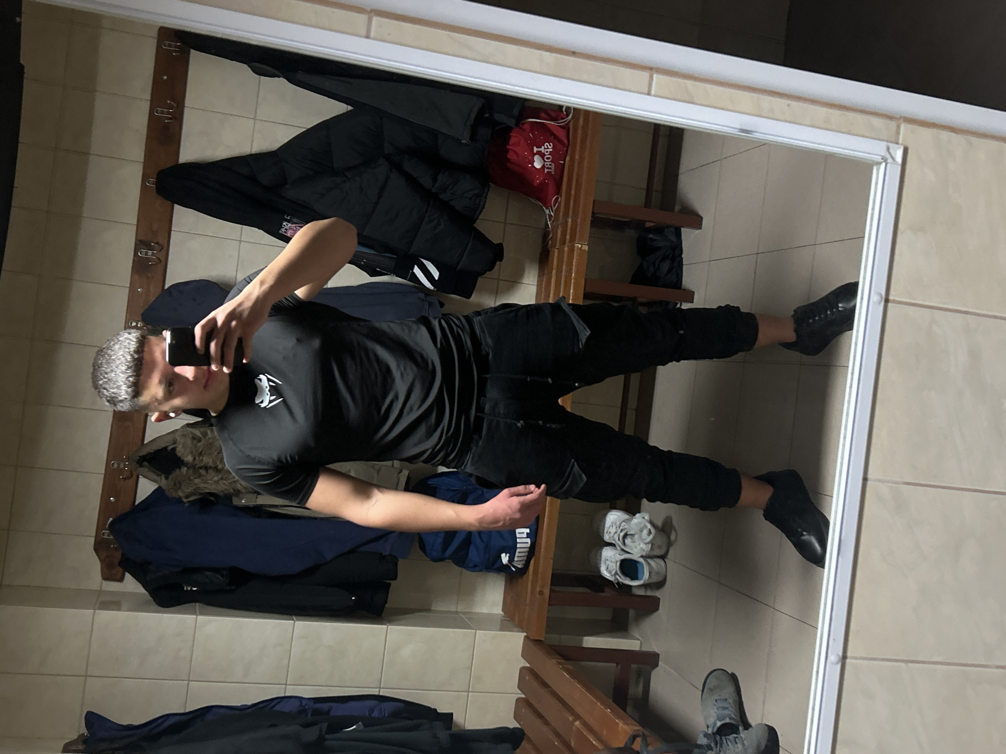 A young man is taking a selfie in a mirror in what appears to be a locker room. He is wearing a black t-shirt, black pants, and black shoes. He is holding a smartphone in his left hand.
Behind him in the reflection are:
* Coat hooks with various jackets hanging on them
* Wooden benches
* Shoes, including sneakers and boots.
* A red bag
The floor and walls are tiled.
The locker room could be located in Zlatograd, Bulgaria, but this is not verifiable from the image.