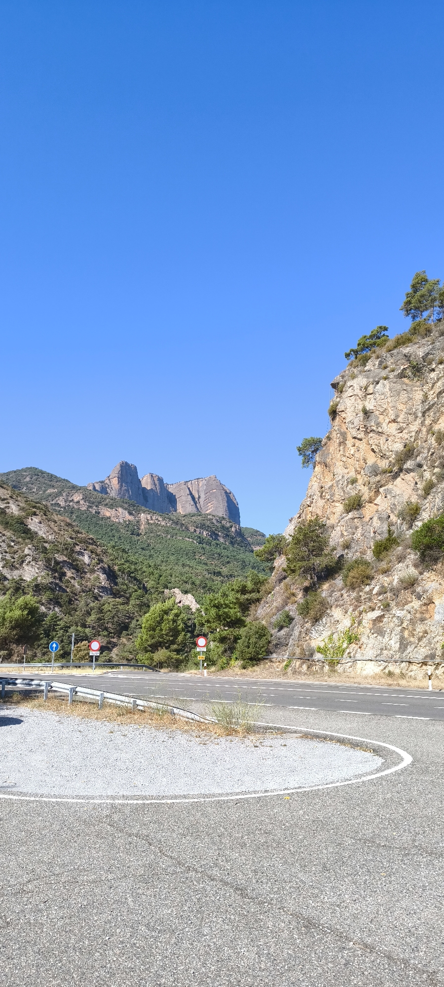 The image depicts a scenic view of a road curving through a mountainous landscape under a clear blue sky. In the foreground, a paved road with white lane markings forms a gentle curve. To the left of the road, a gravel shoulder is visible, bordered by a metal guardrail. Several traffic signs are present along the roadside, including a blue directional arrow sign and two circular red and white prohibition signs.
The midground features a dense forest of green trees climbing the slopes of the mountains. The mountains themselves are rugged and rocky, with distinctive peaks forming a dramatic backdrop. One prominent rock formation stands out with its layered striations. To the right, a steep, rocky cliff face rises, dotted with sparse vegetation and a few trees clinging to its surface.
The location is identified as near the town of Peramola, Spain, suggesting a rural or mountainous area within the country. The scene is illuminated by bright daylight, casting clear shadows and highlighting the textures of the rocks and road. No people or animals are visible in the image.