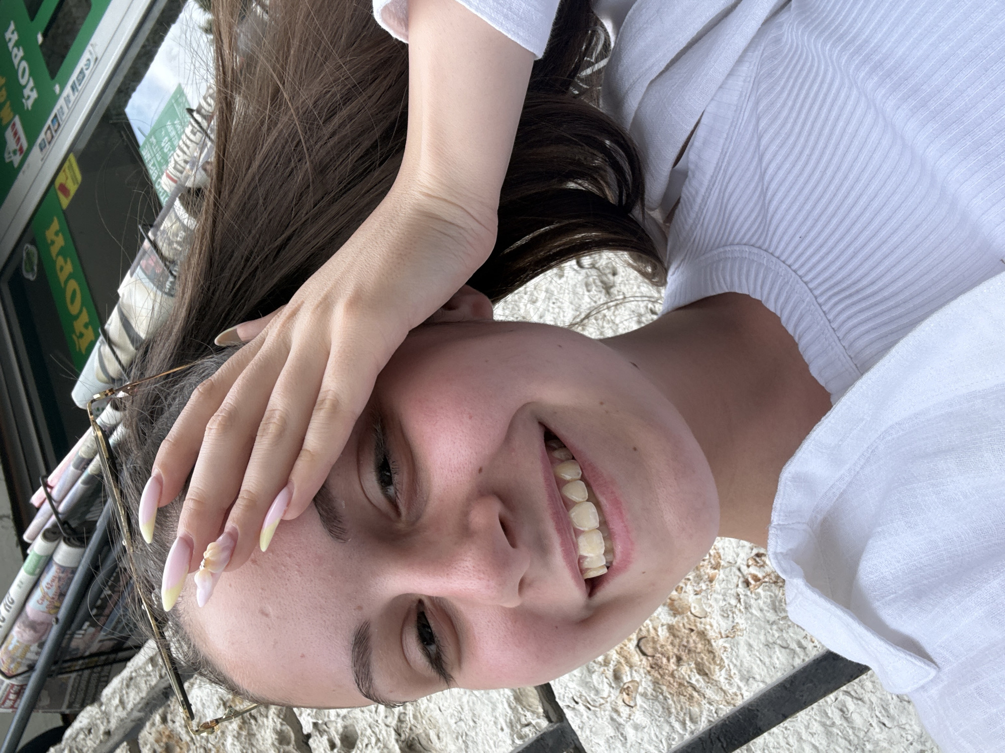 A young woman is taking a selfie outdoors. She has long brown hair and is wearing a white shirt. Her right hand is covering her forehead and eyes. She has long, light pink and yellow French tip nails, with some decorative elements on one nail. She is smiling. In the background, there are newspapers rolled up and tied with string, and a storefront with green signage that appears to read "ИОРИ" (Iori). The wall behind her is made of rough, light-colored stone. The location is the city of Burgas, Bulgaria.