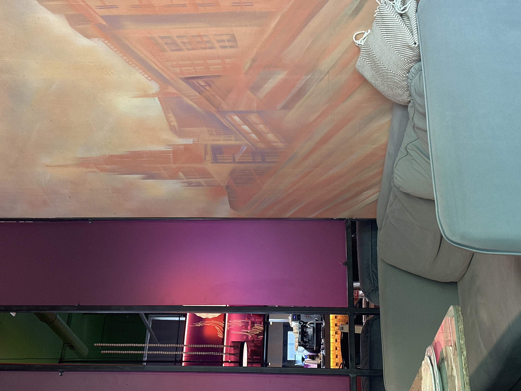 The scene is an interior shot of a room in Novosibirsk, Russia, featuring a wall with an artistic mural of city buildings in shades of brown and orange. Below the mural is a structure with square compartments, illuminated with pink and green lighting. Various objects are visible through the compartments, suggesting a storage or display area. Part of a light-blue sofa with a patterned pillow is positioned on the right side of the frame. The scene appears to be a mix of artistic and functional elements, characteristic of a modern interior space.