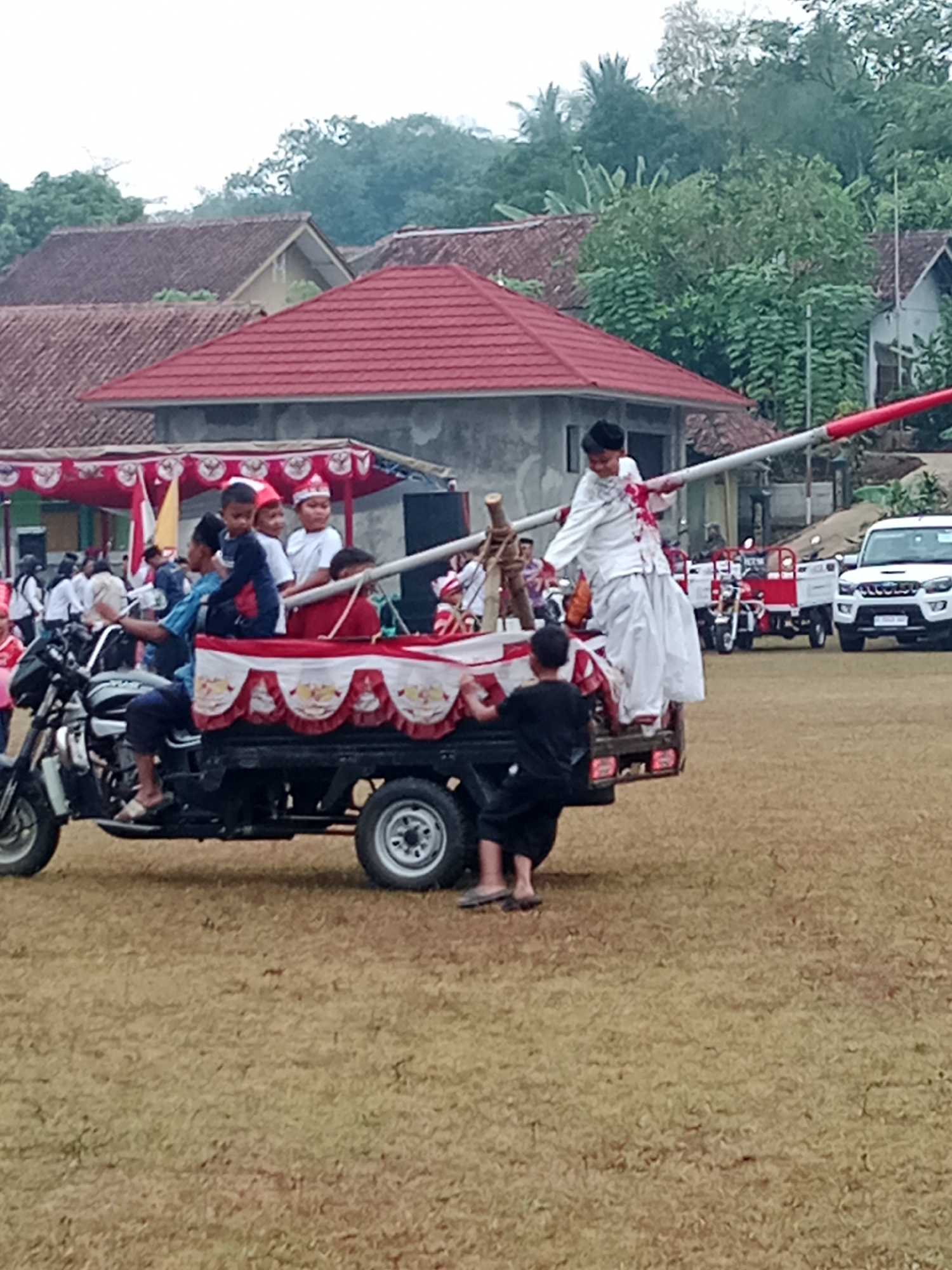 In Bojongmengger, Indonesia, a group of children are participating in a parade. The central focus is a motorized tricycle carrying several young boys dressed in traditional Indonesian attire, including red and white headbands. A boy in a white outfit and headscarf stands on the back of the tricycle, holding onto a long pole adorned with red and white flags, appearing to be performing a balancing act. Another boy in black clothing is positioned at the rear of the tricycle. In the background, other participants in white outfits can be seen, and a white SUV is parked to the right. The scene unfolds on a grassy field, with buildings and trees forming the backdrop under an overcast sky.