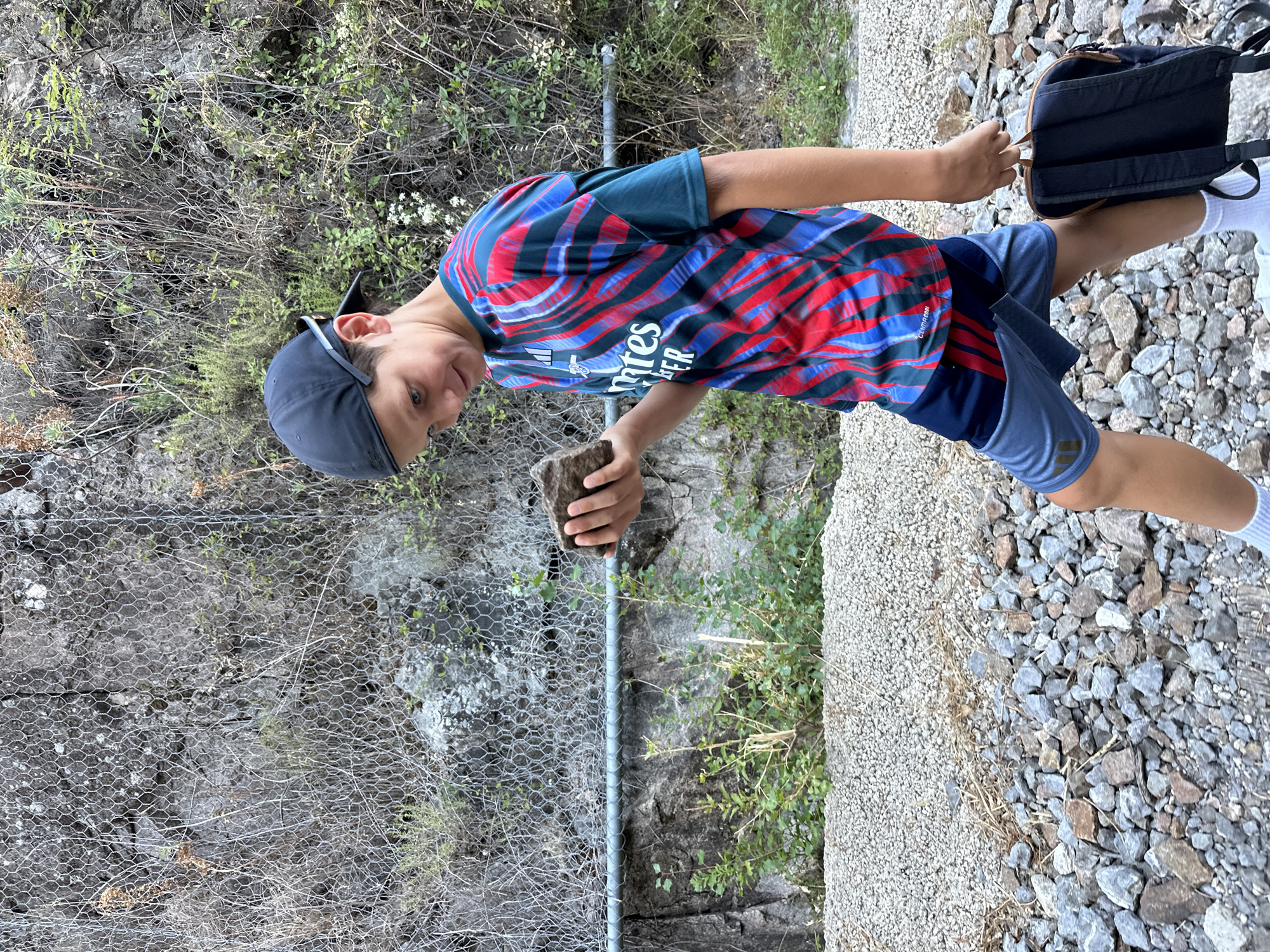 A young boy wearing a baseball cap, a colorful patterned t-shirt, and blue shorts walks along a rocky path. He is holding a rock in his right hand and looking down at it. A black backpack is slung over his left shoulder. Behind him, a rock face is covered with a wire mesh fence, and various plants and trees are growing. The ground is covered with gravel and stones. The scene appears to be outdoors in a natural setting, possibly a hiking trail or mountainous area.