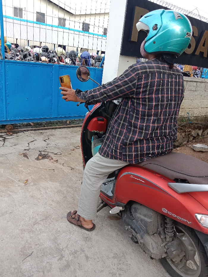 The image shows a man wearing a blue helmet and a plaid shirt, sitting on a red Honda Scoopy motorcycle. He is holding a gold smartphone in his right hand and looking at it. The man is sitting on the motorcycle, which is parked on a paved surface. The motorcycle is facing towards the right side of the image. The man is in front of a blue metal fence with a gate. The fence is in front of a building with a black sign that says "SA". There are many people in the background, but they are blurry and out of focus. They are gathered around the fence and appear to be entering or leaving the building. The scene takes place in Tugu, Indonesia, at coordinates -6.973 latitude and 110.326 longitude. The pavement is cracked and slightly dirty. The motorcycle is clean and the helmet appears to be brand new. The man's left foot is resting on the ground. The man's right foot is resting on the footpeg of the motorcycle. The man is wearing sandals. The scene is captured from a low angle. The focus is on the man and the motorcycle. The image is likely taken by the man, as he is holding a smartphone.