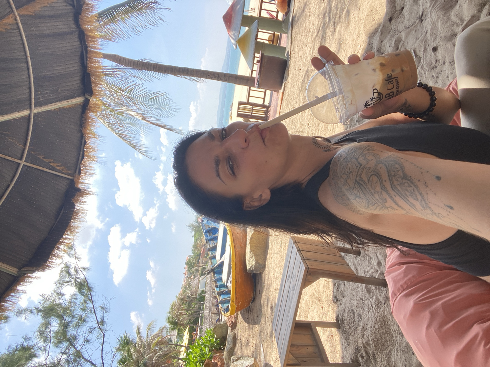 A woman with dark hair and tattoos on her arms is sitting on a pink beanbag on a sandy beach in Phu Thuy Ward, Vietnam. She is holding a clear plastic cup with a brown beverage and a straw, and she is drinking from it. In the background, there are palm trees, beach umbrellas, a wooden table and bench, a yellow and blue boat, and structures with mushroom-shaped roofs. The sky is blue with scattered clouds, and the ocean is visible in the distance.
