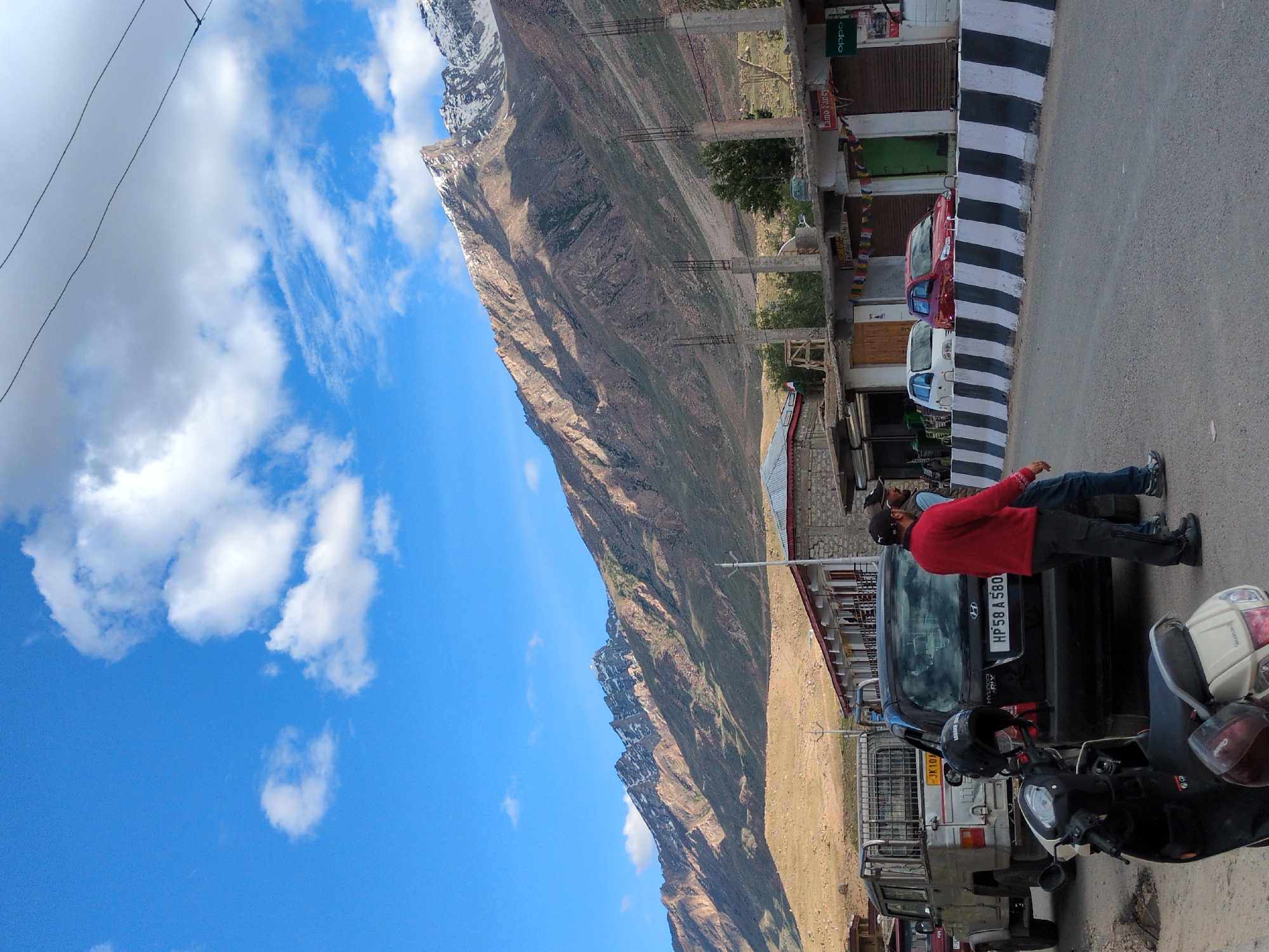 In the city of Padum, India, a mountain landscape is visible with snow-capped peaks under a blue sky dotted with white clouds. In the foreground, a road runs alongside a row of buildings. Two men are standing on the road near several vehicles, including a silver pickup truck, a black car, and a scooter. A white car and a red car are parked in front of the buildings. The buildings appear to be shops, with signs indicating "Lamo Fancy" and "Oppo." The scene suggests a street in a town situated in a mountainous region.