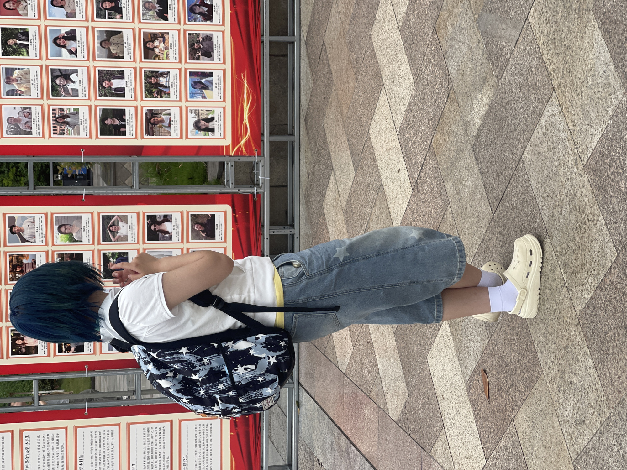 A medium shot captures a person with blue hair standing in front of a display in what appears to be Chongqing High-tech Zone, China. The display is a red banner featuring multiple small photographs of people. The person, facing away from the camera, wears a white t-shirt, jean shorts with white stars, white socks, and white clogs. They also wear a black and white patterned backpack. The ground is paved with light brown stones in a diagonal pattern. The text on the display is in Chinese.