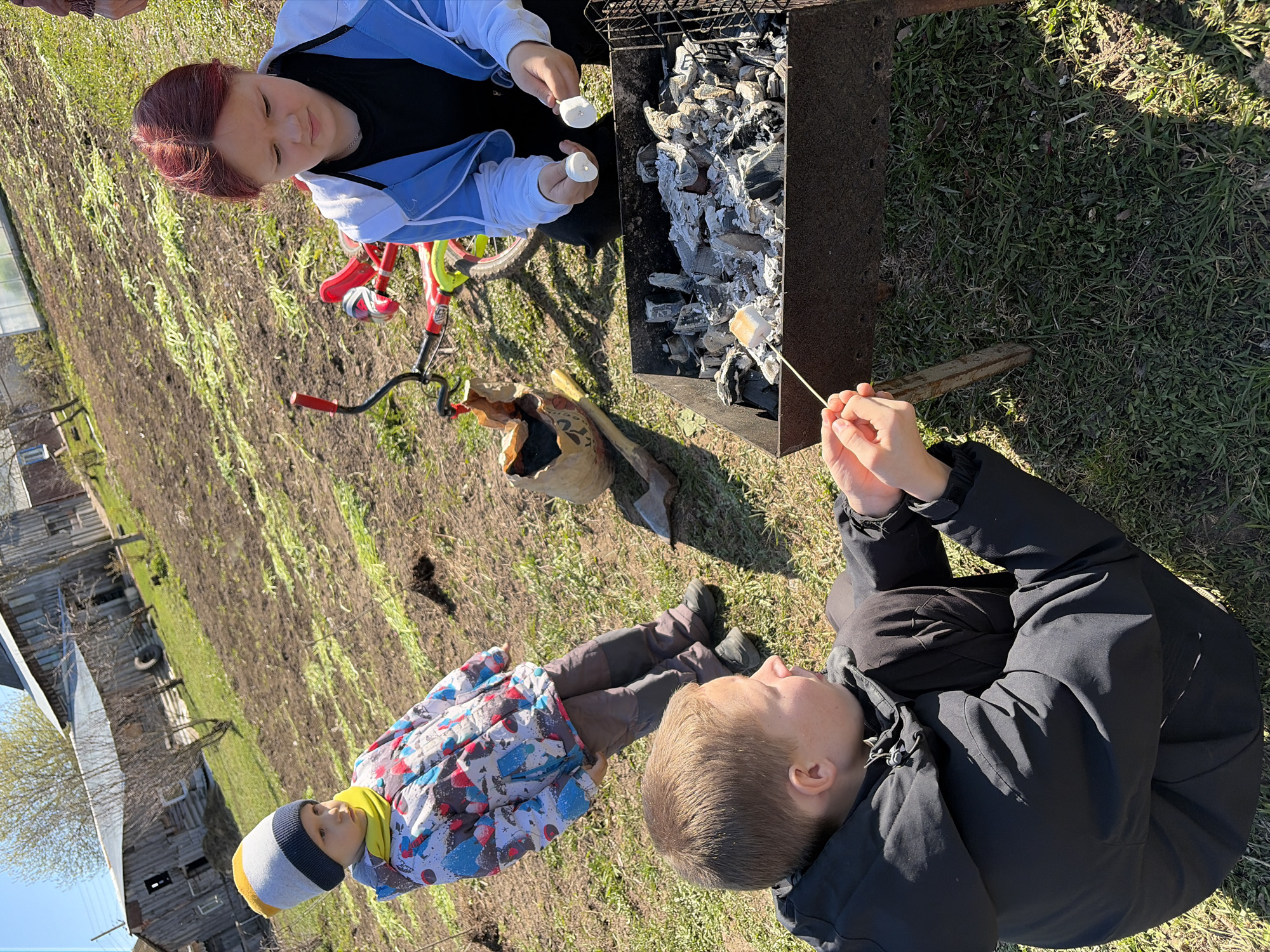A medium, outdoor shot depicts two individuals roasting marshmallows over a grill in a rural environment, likely near the city of Чумна, Russia.
A fair-skinned woman with red hair is seen holding two marshmallows. She is dressed in a black shirt under a light blue and white jacket. Next to her, a child with a fair complexion is holding a marshmallow on a stick over the grill. The child is wearing a black coat. A second child, wearing a colorful jacket and a grey, yellow, and blue beanie, is seated on the grass.
The grill is filled with hot coals. Beside the grill is a small red and green bicycle. A beige bag is lying on the ground near the bicycle.
The setting appears to be a grassy field. In the background, there is a wooden building with a visible tire, suggesting a rural or suburban environment. The scene is bathed in natural sunlight, indicating it is daytime.