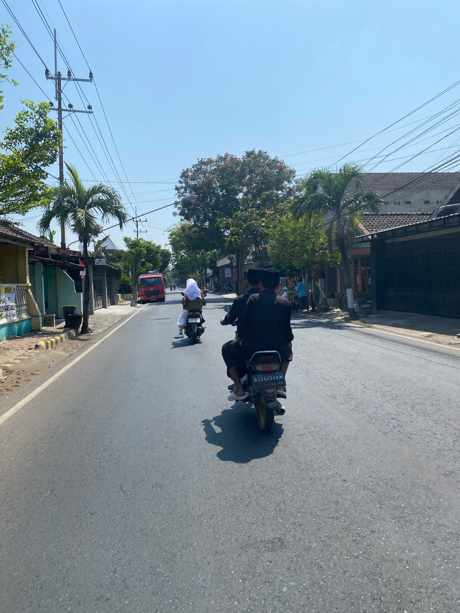 The image depicts a sunny day on a paved road in Kediri City, Indonesia. Two individuals, one on the front and one on the back, are riding a motorcycle, both wearing black clothing and black peci hats. The motorcycle has the license plate "AG5425VAM" and the date "02-25" visible. Ahead of them, another motorcycle carries two people, with the passenger wearing a white hijab. Further down the road, a red truck is visible. The environment includes houses with tiled roofs, palm trees, and overhead power lines. Some shops and a yellow and black striped curb are also present along the side of the road.