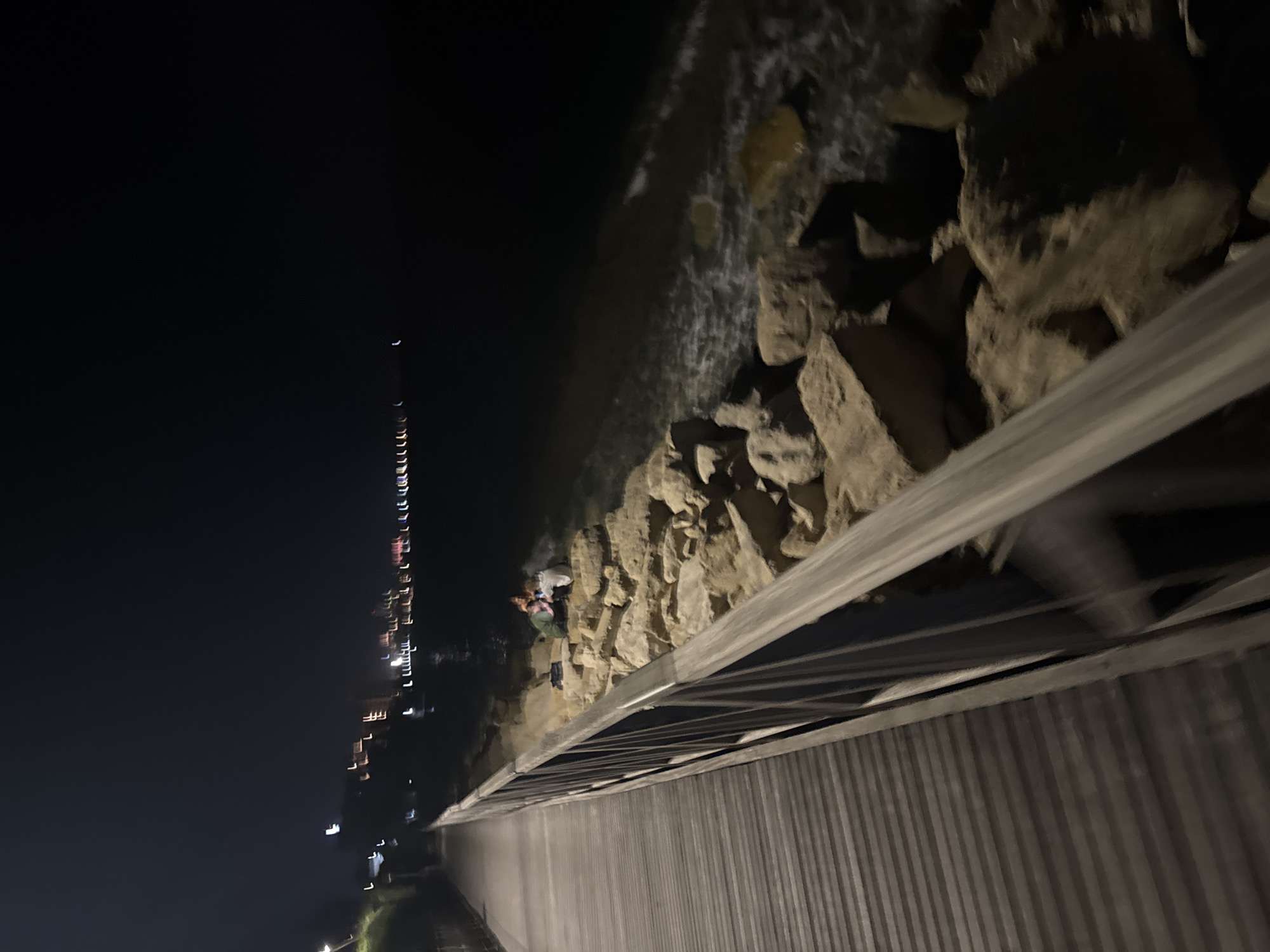 The image depicts a nighttime scene along a waterfront promenade in Agios Tychona Community, Cyprus. A wooden walkway with a metal railing extends into the foreground, running diagonally from the lower left to the upper left of the frame. To the right of the walkway, a rocky embankment borders the dark water. On the embankment, three figures are seated among the rocks, appearing to be engaged in conversation or activity. In the distance, across the water, city lights form a blurred horizontal line, indicating an urban or developed coastal area. The sky is dark and clear, with no visible stars. The water shows some movement, with small waves lapping against the rocks. The overall impression is a tranquil evening by the sea.