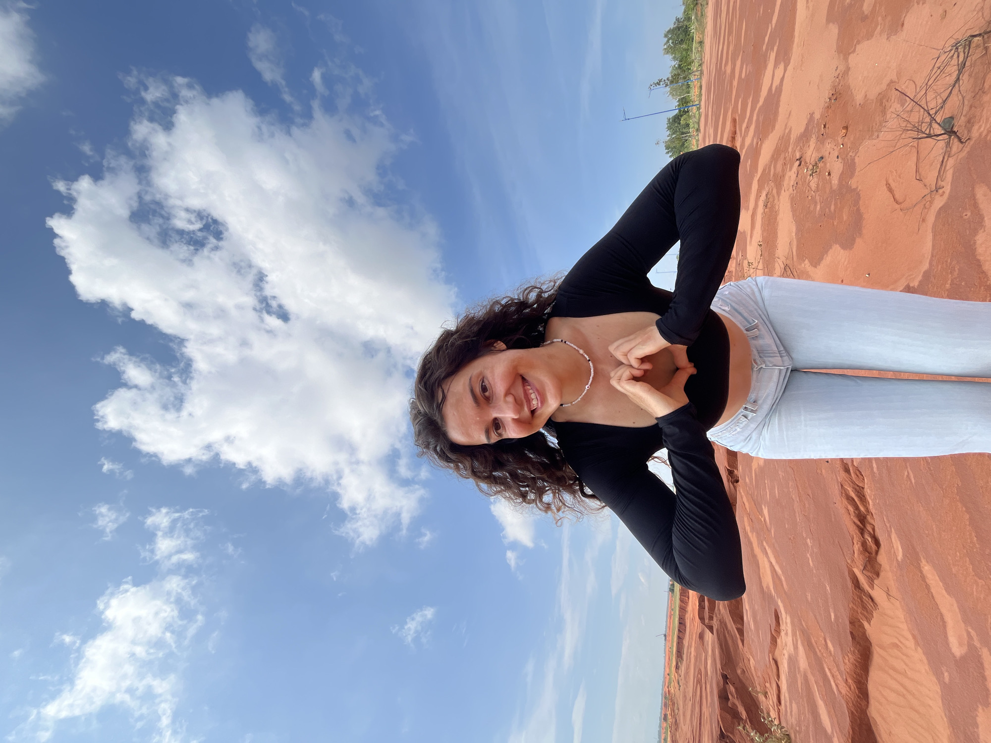 A young woman with dark, curly hair smiles at the camera, her hands forming a heart shape in front of her chest. She is wearing a black long-sleeved crop top and light-wash jeans. The background features a vast expanse of red, sandy terrain, characteristic of the Red Sand Dunes in Mui Ne, Vietnam. The sky above is a bright blue, dotted with fluffy white clouds. In the distance, scattered trees and utility poles are visible, hinting at the edge of the dunes.