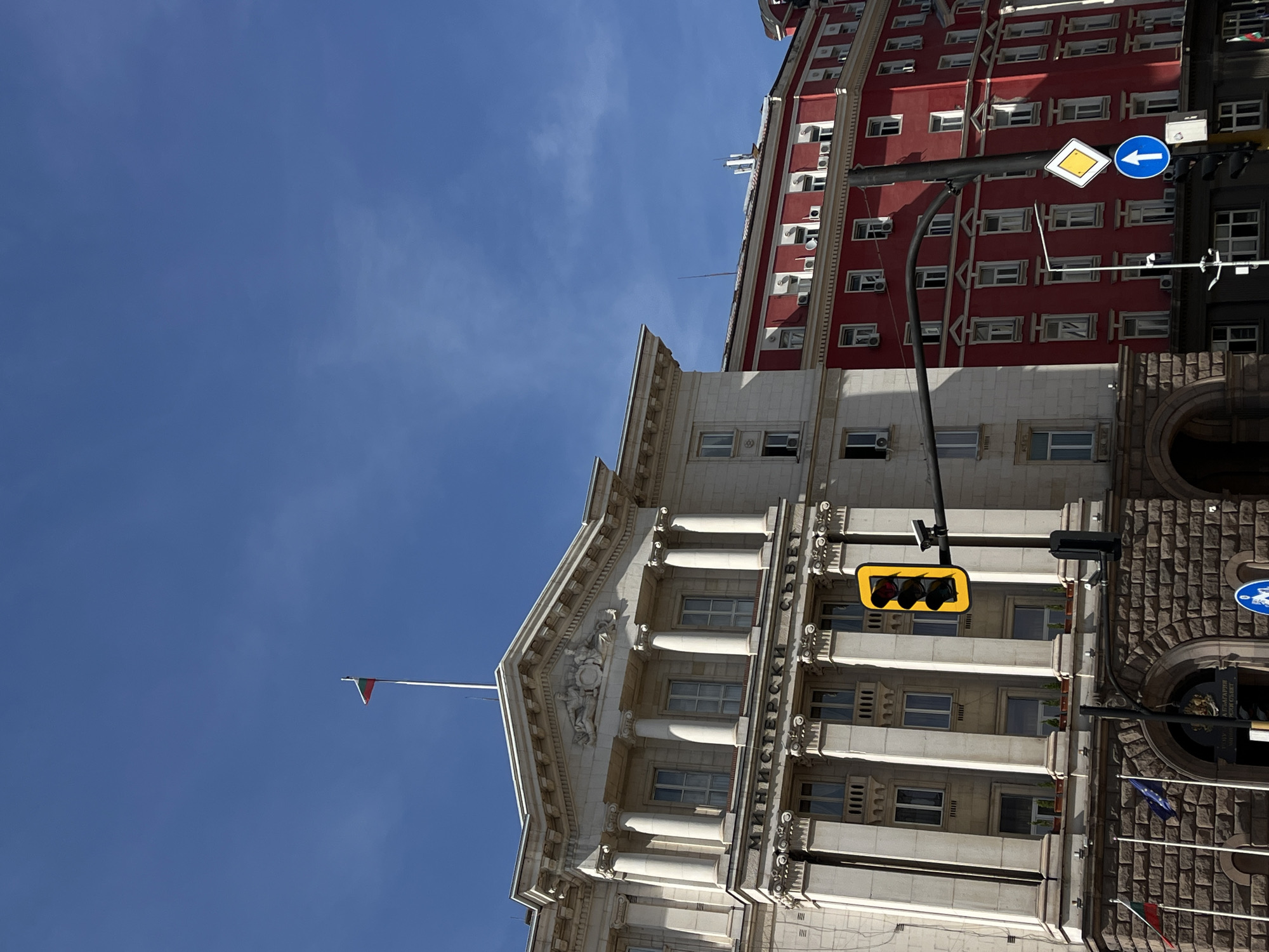 The image depicts two buildings in Sofia, Bulgaria, against a clear blue sky. The first building, constructed from light-colored stone, is ornately decorated with columns and classical architectural elements. A flagpole with a small flag extends from its roof. To the right, a darker, red-brick building displays numerous windows and air conditioning units. A traffic light hangs in the foreground, displaying a yellow light. Road signs are attached to a pole, including a blue sign with a white arrow and a yellow diamond sign. Flags and building signs are visible in front of the stone building. The image is oriented at an oblique angle.