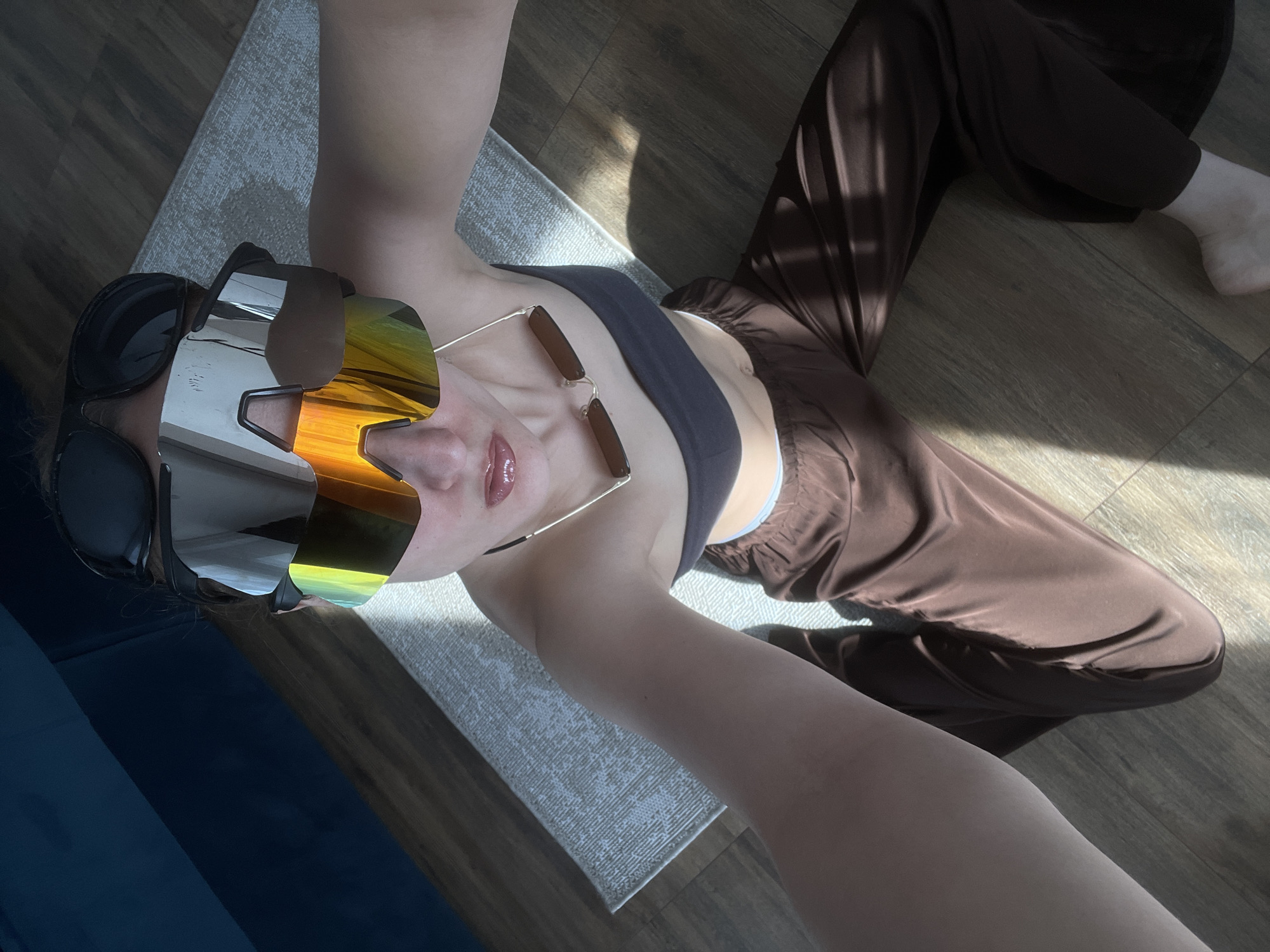 A woman is lying on the floor, wearing a gray sports bra and brown satin pants. She is also wearing two pairs of sunglasses, one on top of the other. One pair is black and the other has silver and gold mirrored lenses. She is lying on a gray rug. The floor is wooden. The woman's right leg is outstretched towards the upper right corner of the image, with her foot near the corner. Part of a blue surface is visible in the lower left corner. Sunlight is streaming in from the upper right, creating shadows on the pants. The location is indoors, likely a home, but there is no information in the image that indicates it is in Подшивалово, Russia.