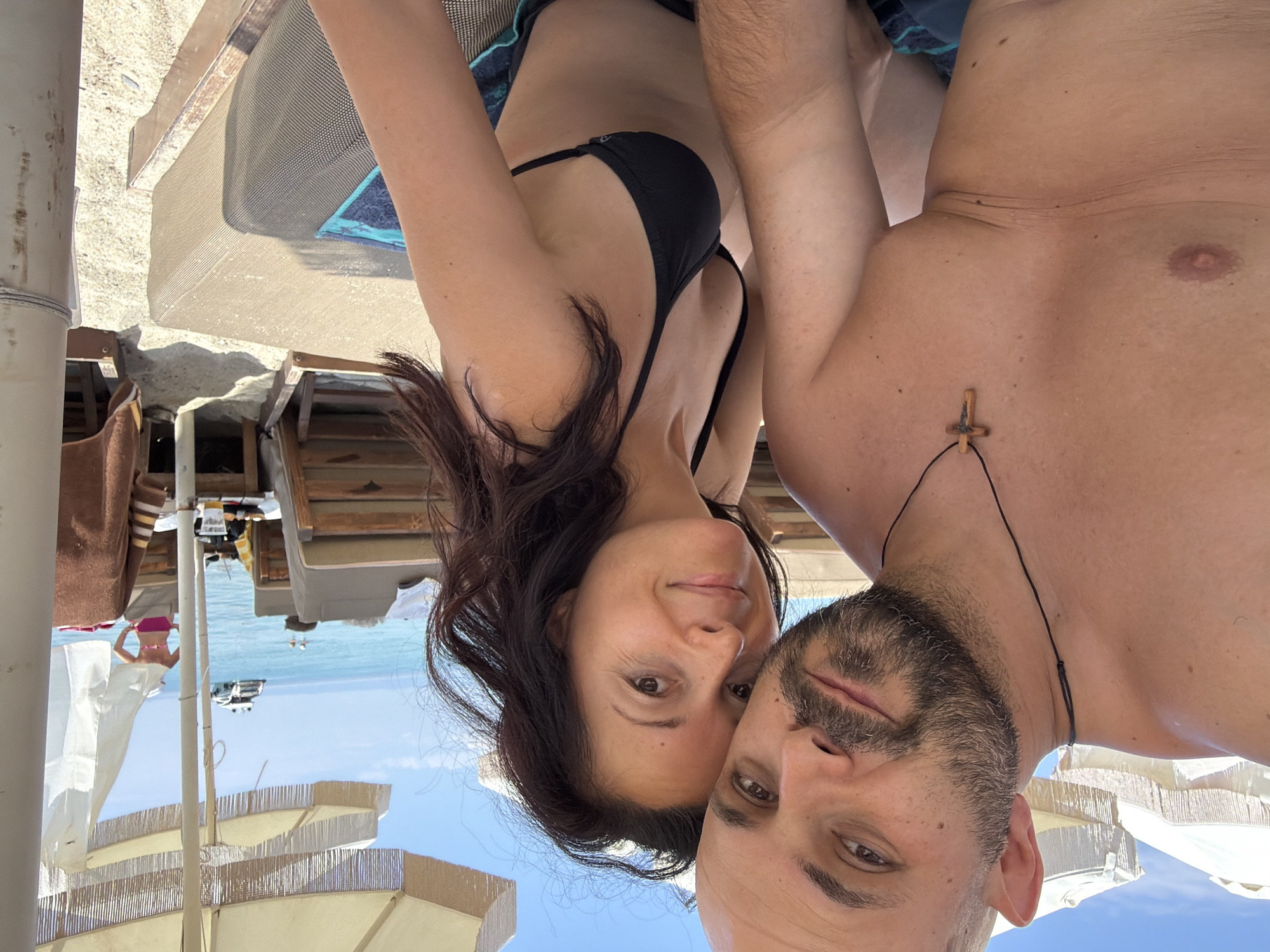 The original image is a selfie taken by a couple on a beach. The man on the left has a shaved head and a beard, wearing a black necklace with a wooden cross. The woman on the right has dark, long hair and is wearing a black bikini top. They are both smiling and looking at the camera. Behind them, beach chairs and umbrellas are visible, as well as the turquoise sea and a white yacht in the distance. The scene suggests a relaxed holiday atmosphere. The location appears to be a beach in the Pallini Municipal Unit of Greece, likely a popular tourist area given the presence of beach amenities and a yacht.