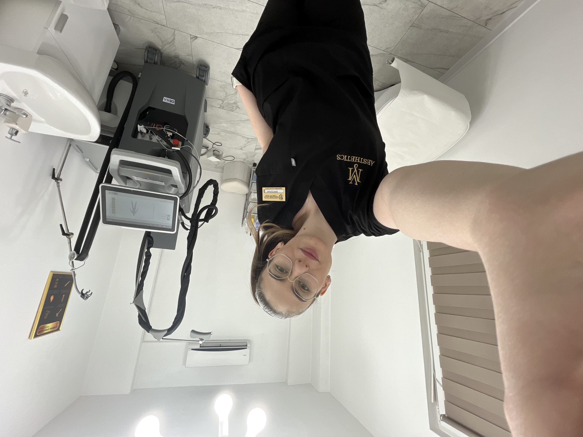 The image shows a room with medical equipment, including a DEKA device. A woman wearing a black scrub suit with the "AESTHETICS" logo is visible. The location is Unknown, Unknown.