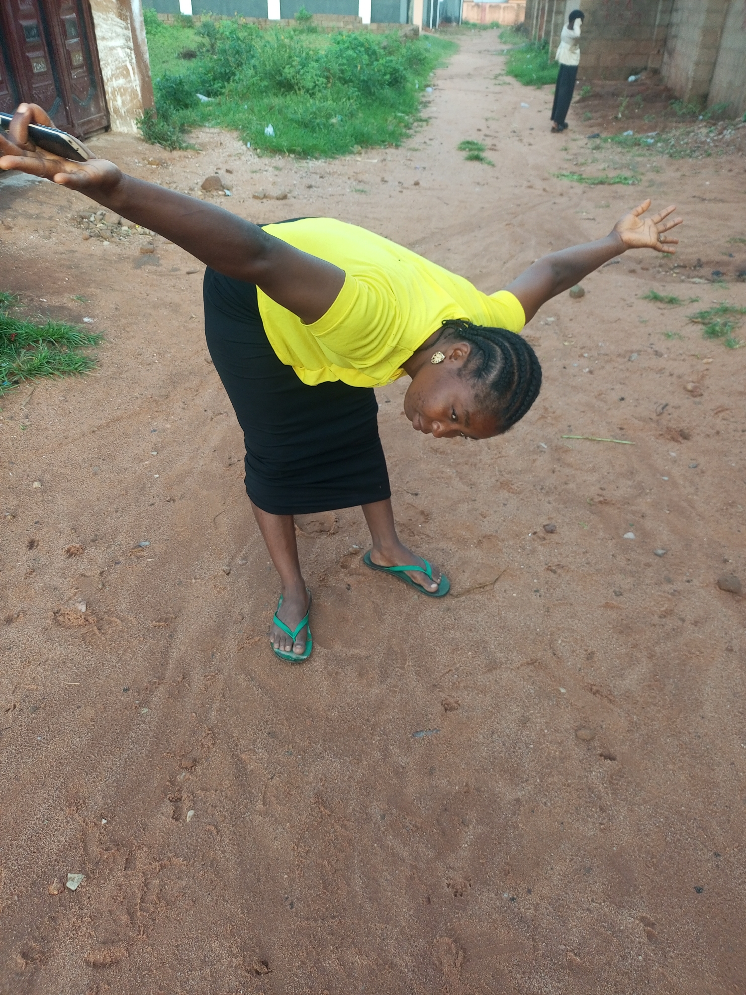 In Tempure, Nigeria, a woman in a bright yellow shirt and a black skirt is captured mid-motion on a sandy path, leaning forward with her arms outstretched as if about to pick something up or greet someone. She is wearing green flip-flops. In the background, a person in a light-colored shirt and dark pants stands further down the path, facing away from the camera. The environment appears to be an unpaved road with sparse vegetation on either side and a low wall in the distance.