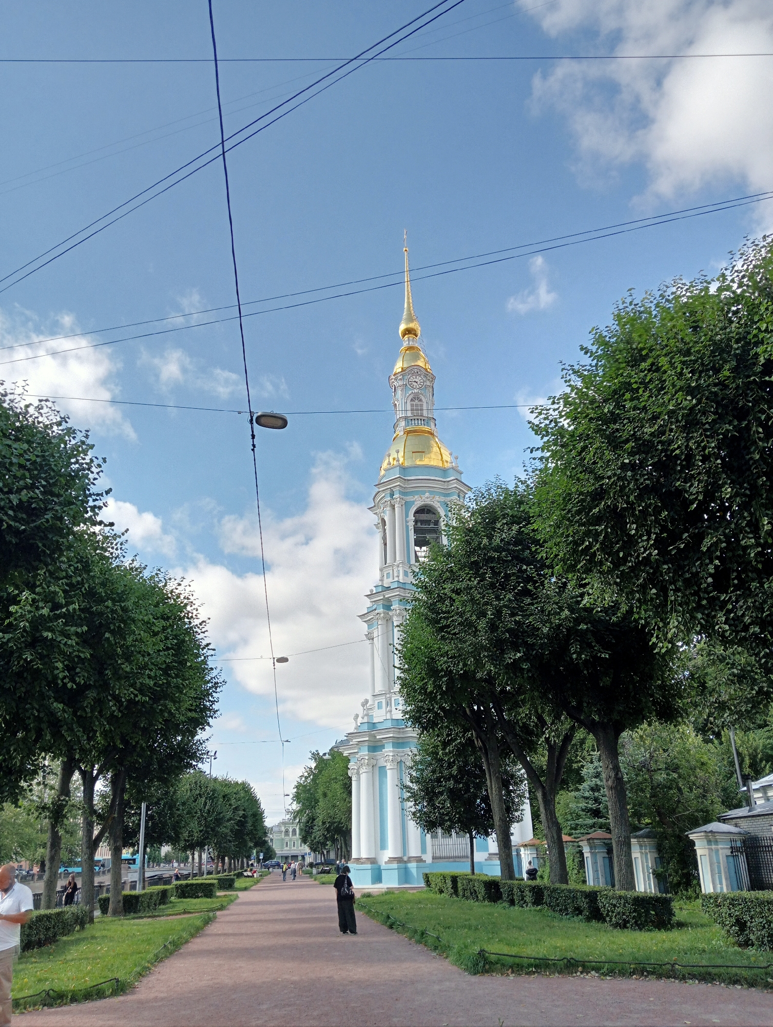 This is a vertical, medium shot photograph taken on a sunny day in Saint Petersburg, Russia. The image shows a wide, paved walkway lined with manicured green hedges and trees. In the background, a tall, slender bell tower with a golden spire dominates the skyline. The tower is white with blue accents and features a clock face. To the right of the walkway, a portion of a building with white columns and blue trim is visible, consistent with St. Nicholas Naval Cathedral, a prominent landmark in the city. Several people are present, including a man in a white shirt and tan pants on the left, a person in dark clothing walking away from the camera in the center, and other figures further down the path. Overhead, multiple power lines crisscross the bright blue sky, which is dotted with white clouds. The overall scene depicts a peaceful urban park setting with a significant religious structure.