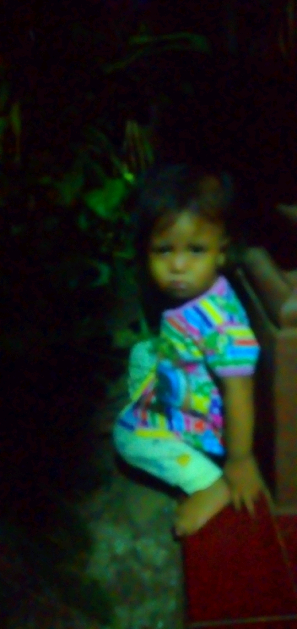 A blurry, low-light image depicts a young child sitting on a dark red surface, possibly a step or a mat. The child is wearing a colorful, striped shirt with horizontal bands of green, yellow, blue, pink, and white. The child's lower body is covered by white pants with yellow designs. The background is dark and out of focus, with some blurred green foliage suggesting an outdoor or dimly lit environment. The child's face is partially visible, with a neutral expression. The overall scene appears to be captured in Rahong, Indonesia, given the context of the request, but no specific city landmarks are identifiable within the image itself. The lighting suggests it may be nighttime or dusk, and the image quality is poor due to motion blur and low resolution.